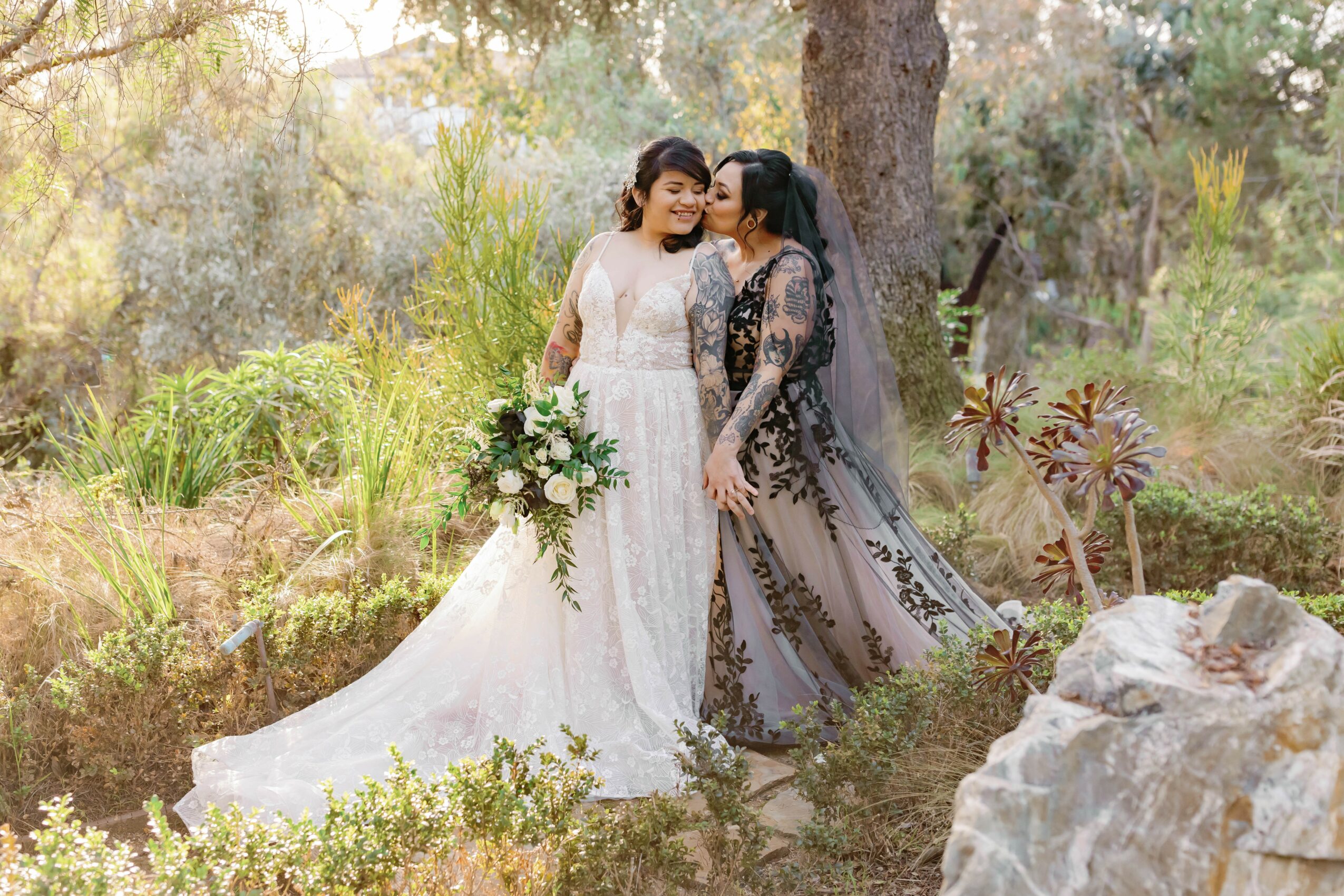 Two women in wedding dresses stand outdoors, one holding a bouquet, surrounded by trees and greenery.