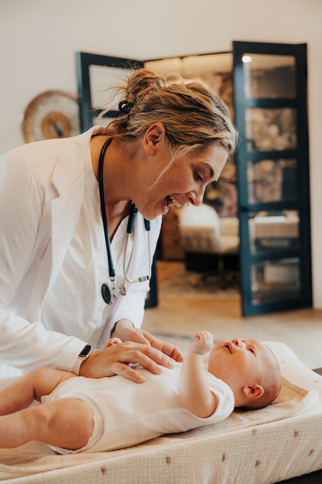 A woman in a white coat interacts with a baby lying on a changing table, smiling and engaging with the infant.