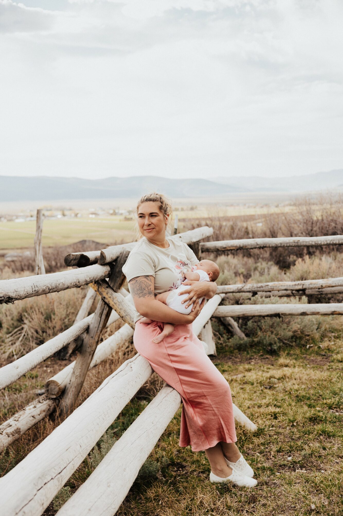 Woman sitting on a wooden fence holding a baby outdoors with fields and sky in background.