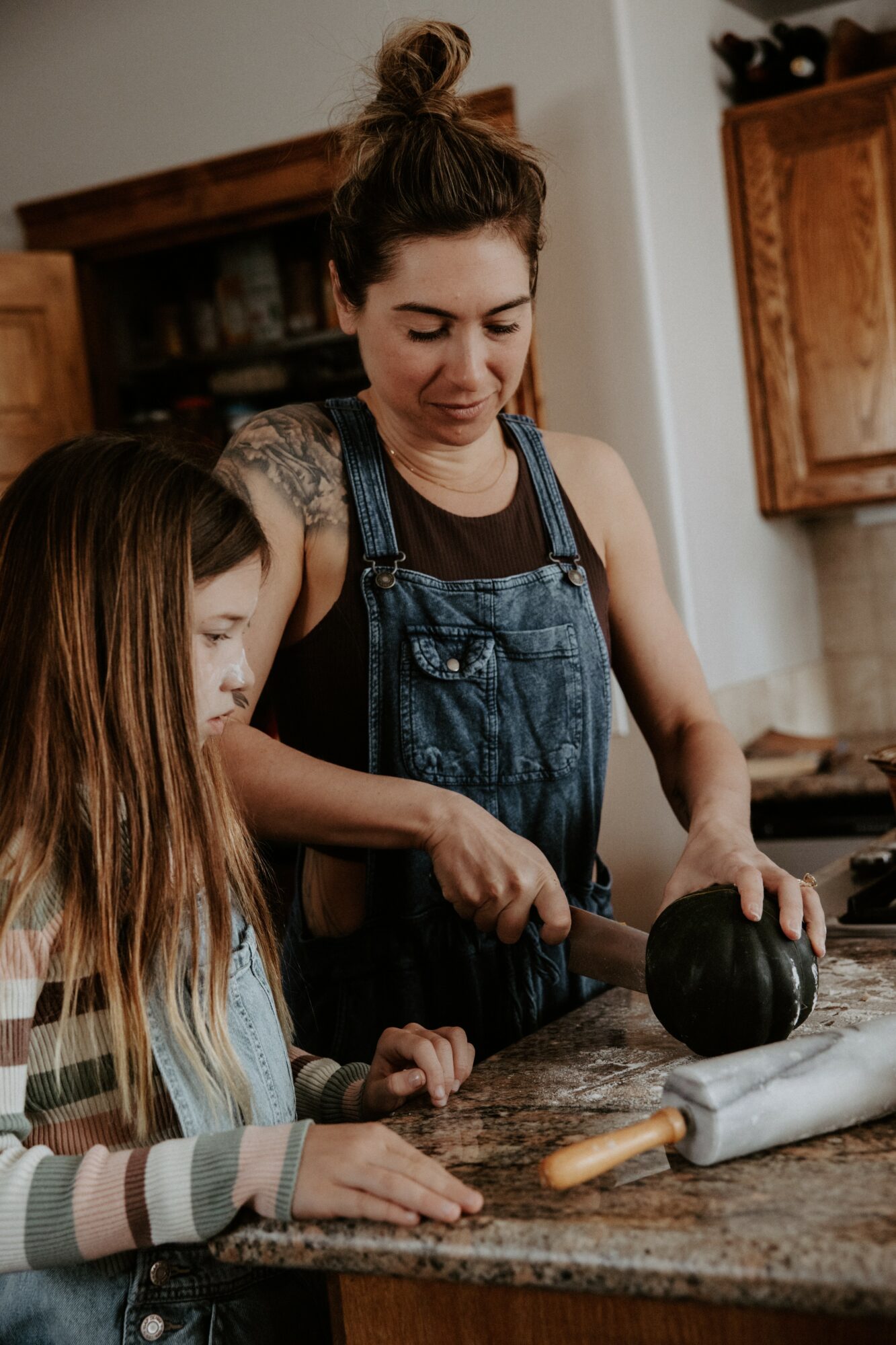 Woman and girl baking together in kitchen, woman holding rolling pin, girl watching, kitchen cabinets in background.