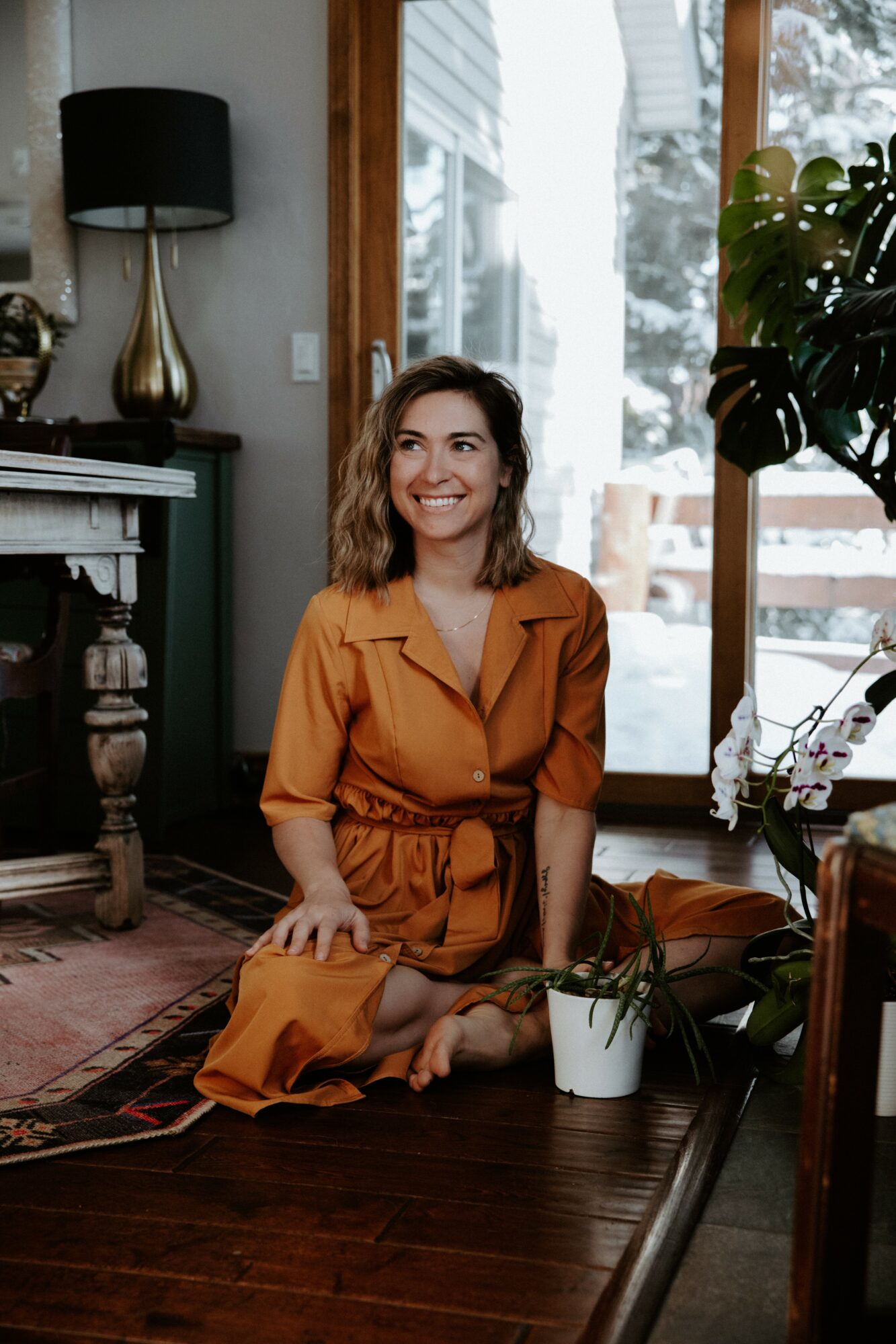 Woman sitting on the floor smiling, with a plant in a white pot nearby, in a room with large windows and wooden furniture.