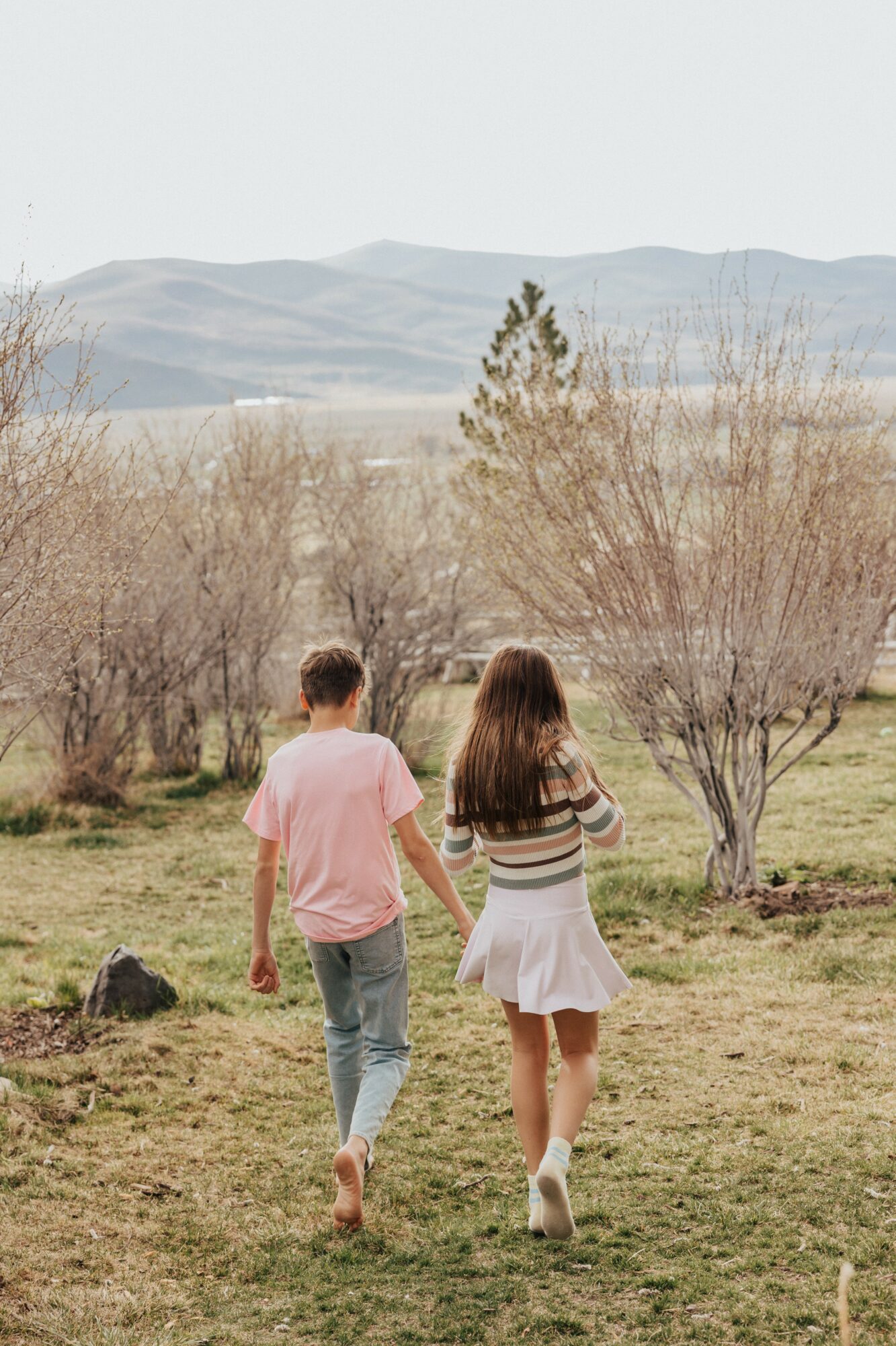 Two children, a boy and a girl, walk hand in hand in a grassy field with trees and mountains in the background.
