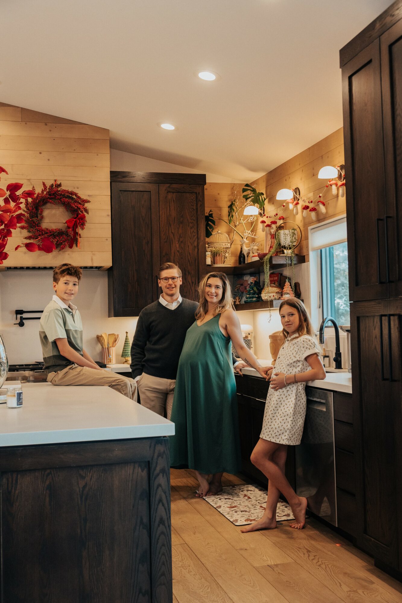 Four people in a kitchen, two women and two children, standing near a window with warm lighting and decorations.