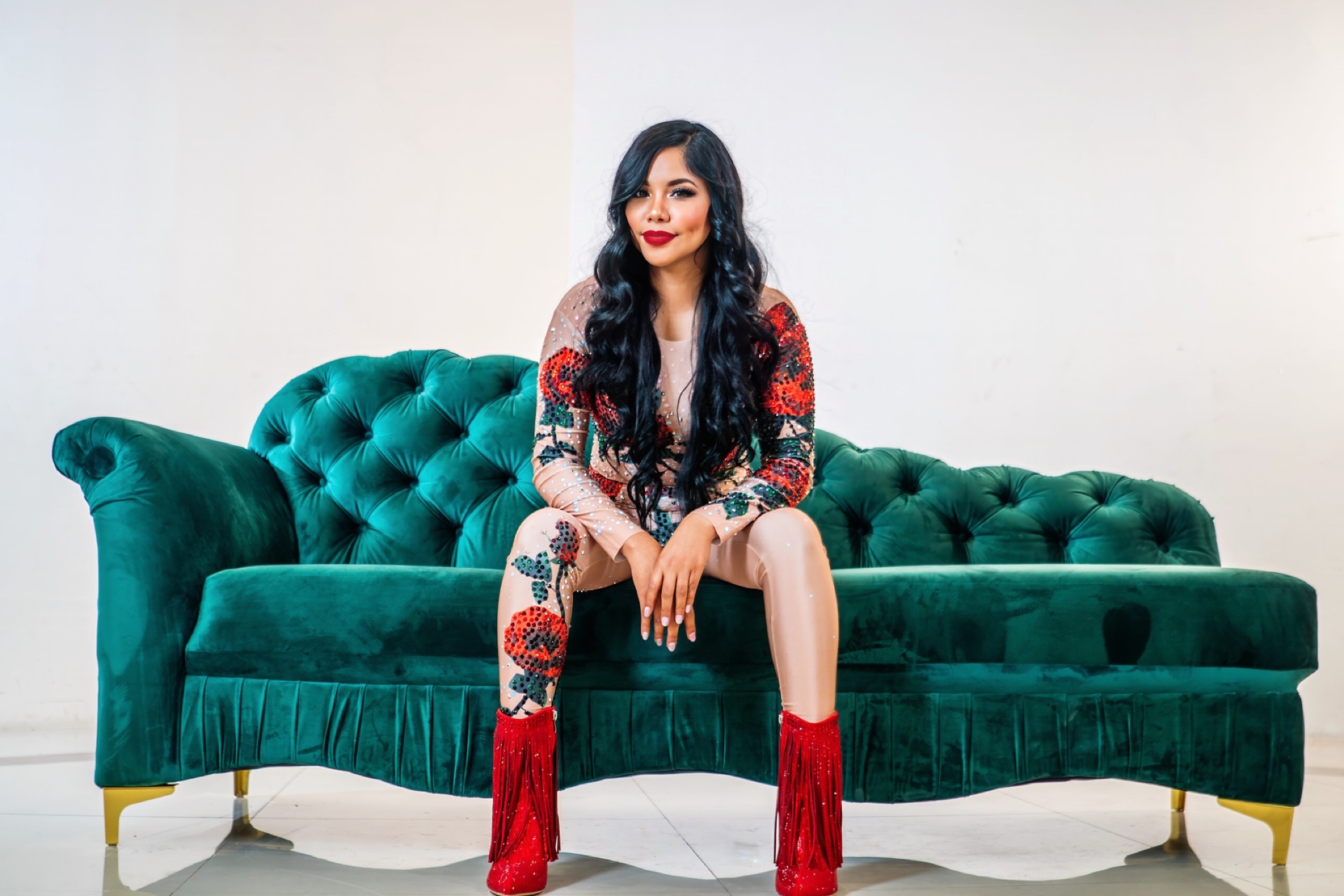 Woman with long dark hair and red lipstick sitting on a green velvet sofa, wearing a floral outfit with red fringed boots.
