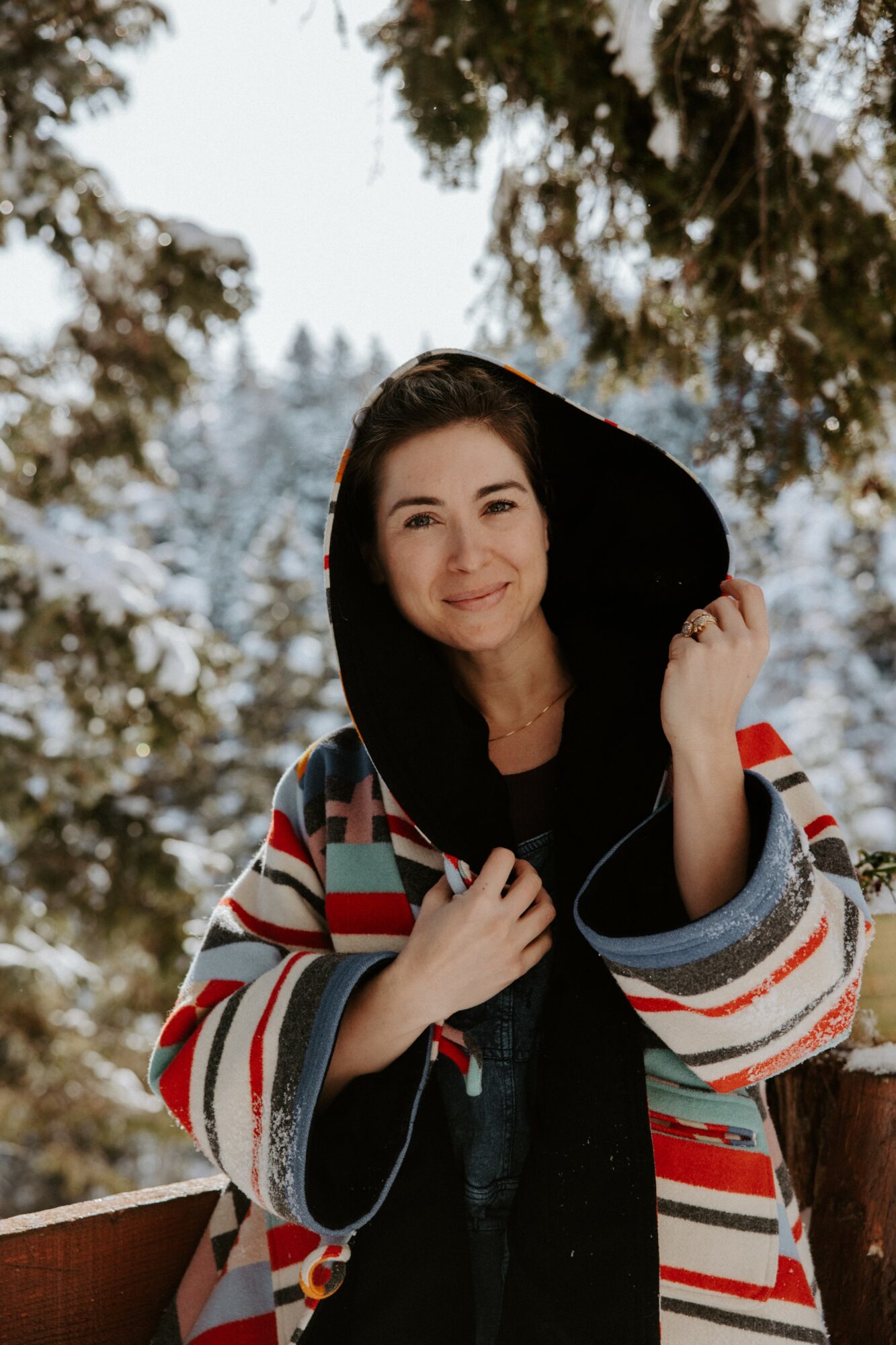Young woman outdoors in winter, smiling, wearing a striped jacket and holding a hood, trees and snow in background.