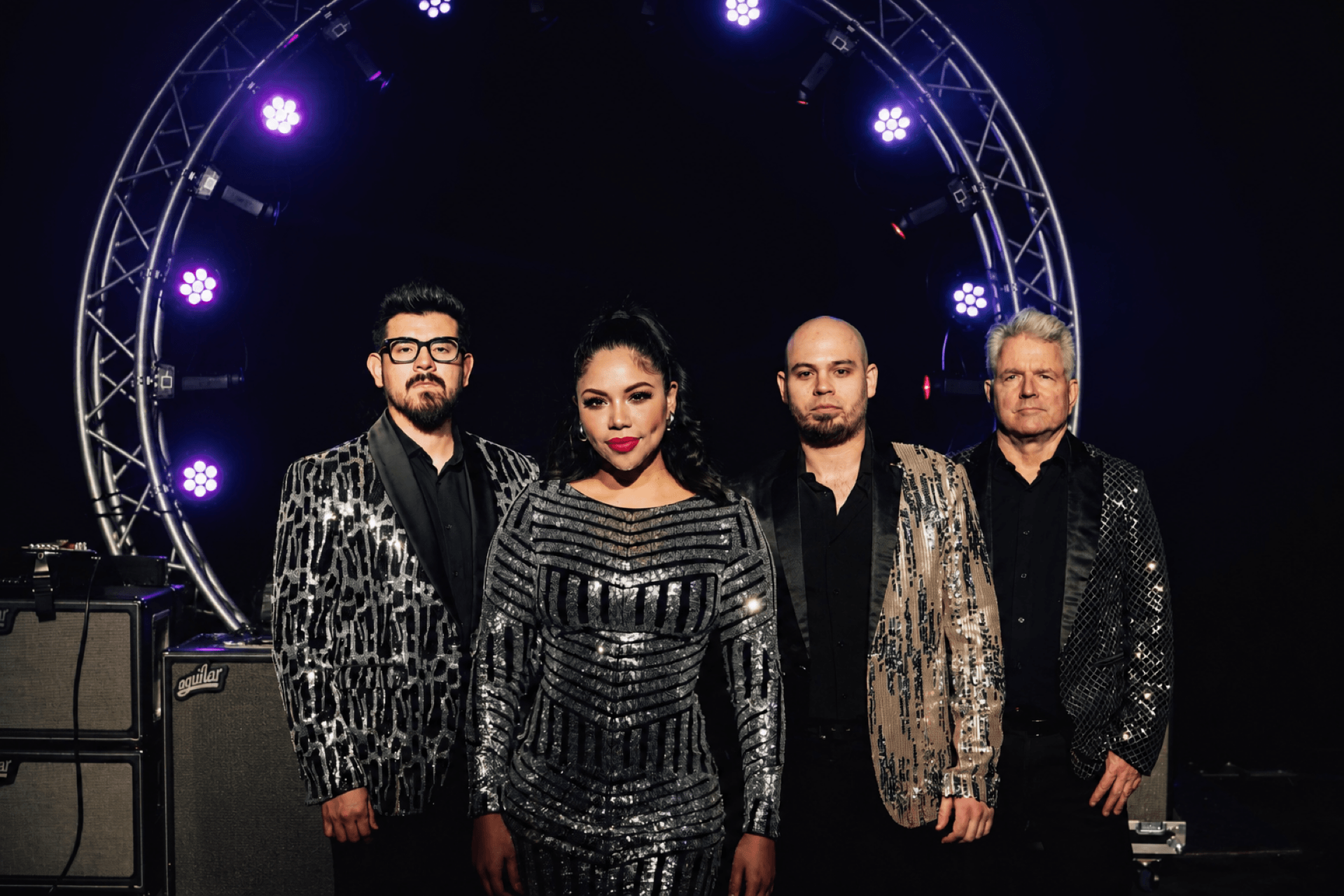 Four people stand in front of a circular light structure on a dark stage, dressed in shiny, patterned clothing.