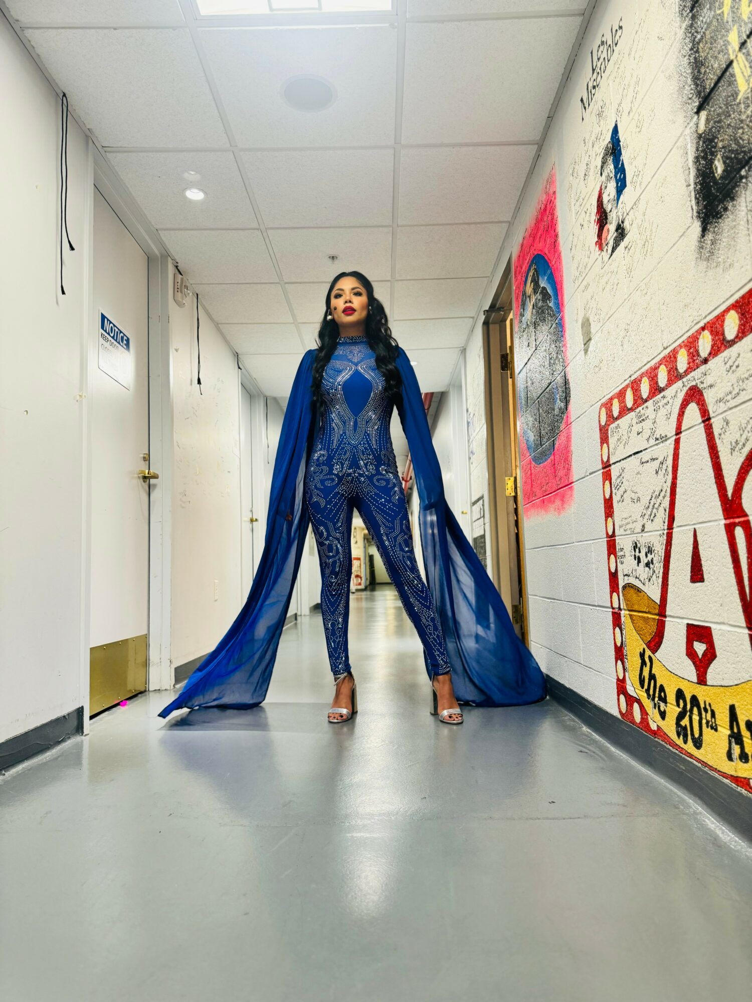 Woman in blue outfit standing in hallway with colorful wall art, wearing high heels and a flowing cape.
