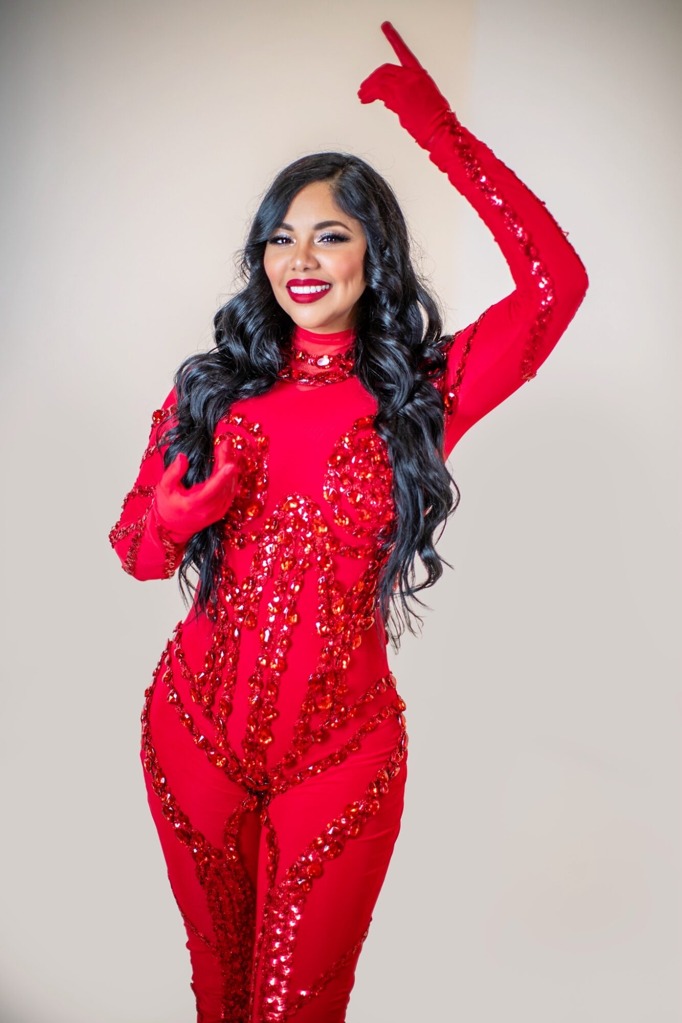 Woman in a red sequined dress with long black hair, smiling, raising her right arm, against a plain background.