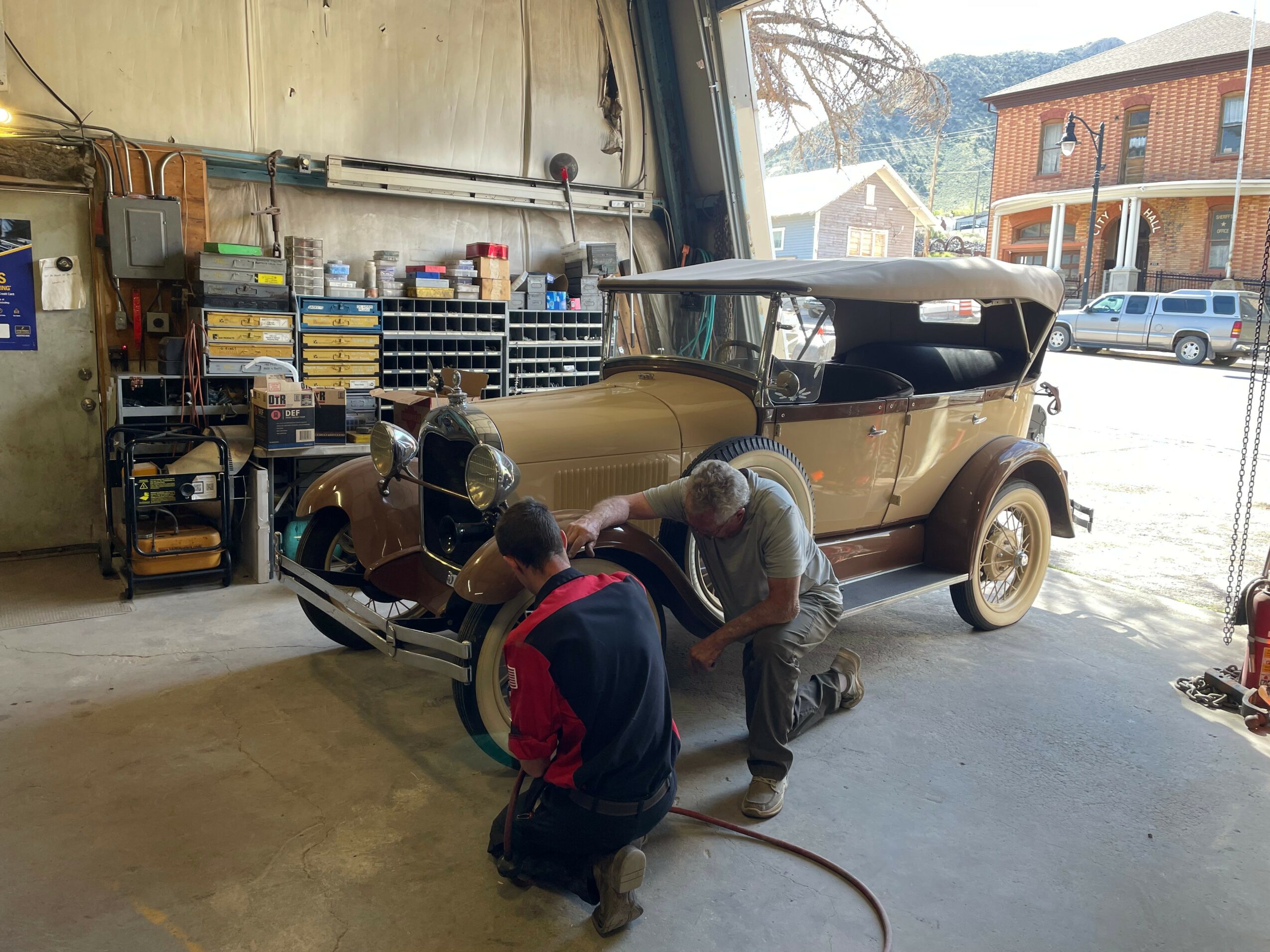 Three people working on a vintage car inside a garage, with tools and shelves in the background.