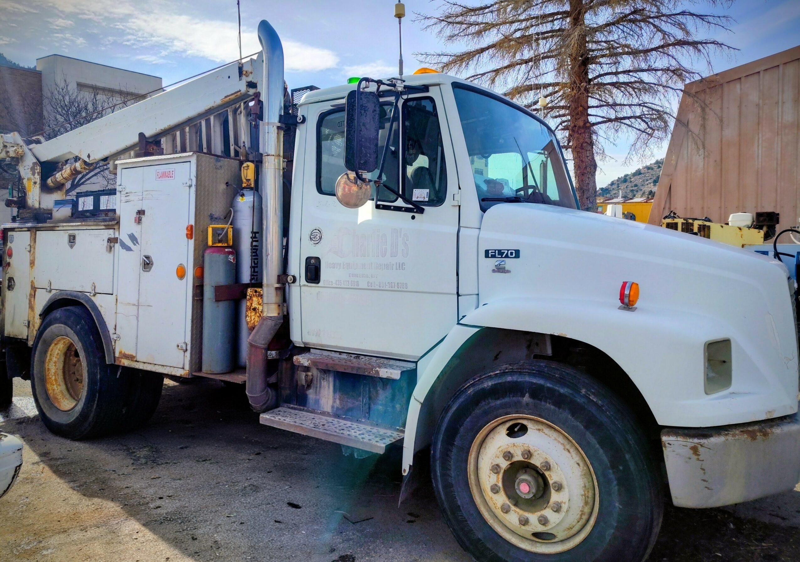 Utility truck with equipment, parked outdoors with trees and buildings in background.