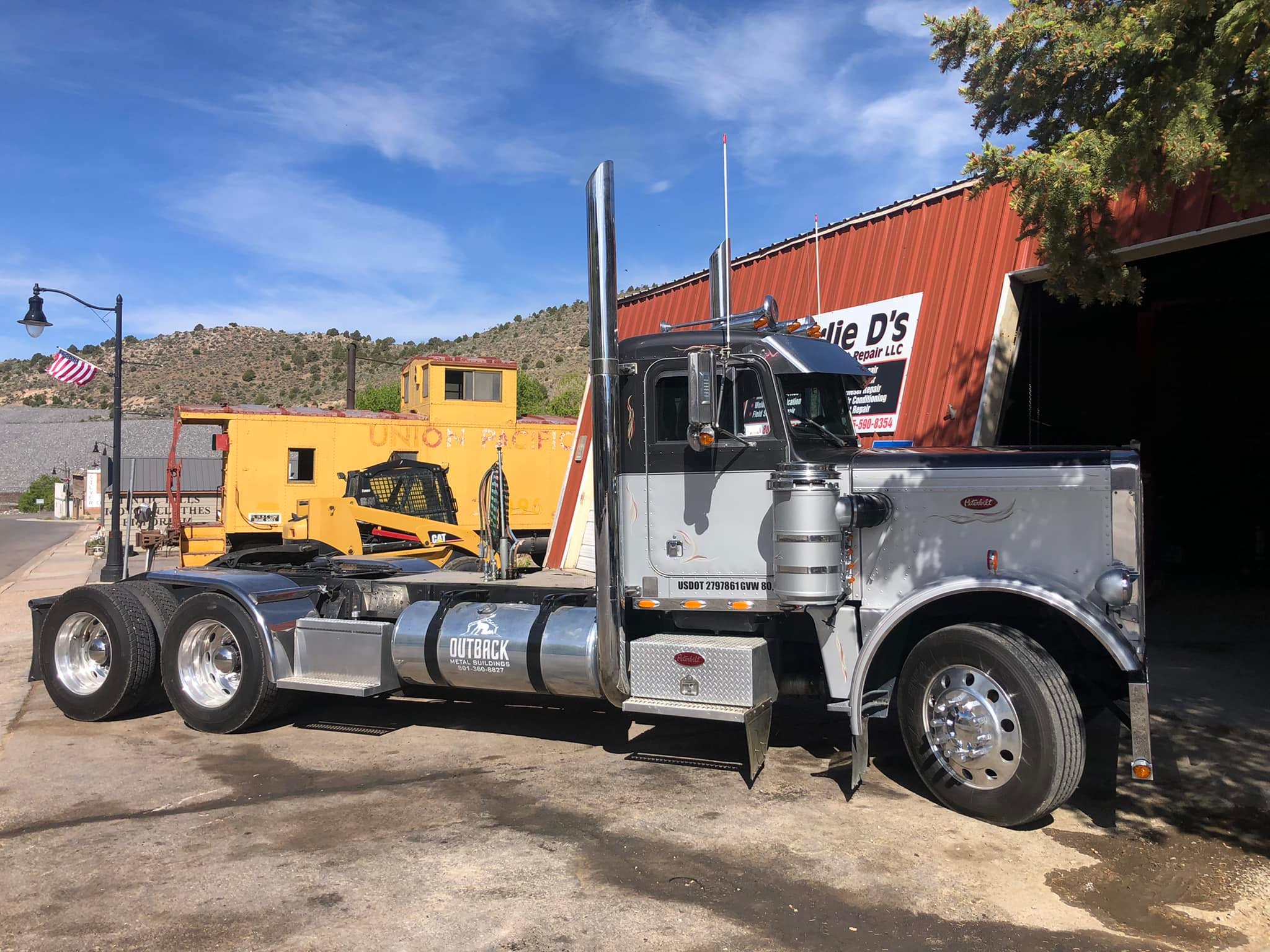Semi-truck parked outside a building with a red roof, yellow building, and a landscape with hills and trees in the background.