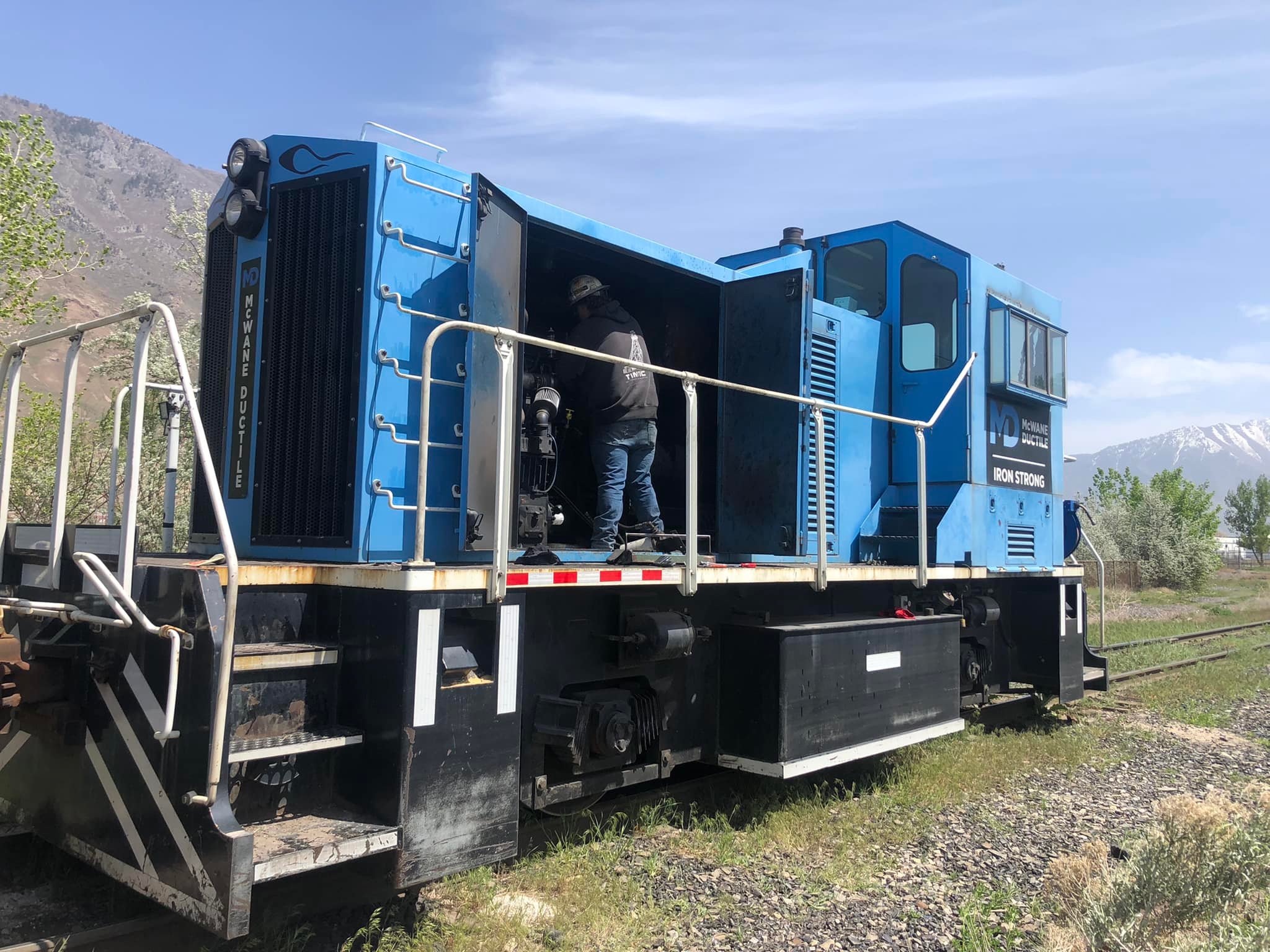 Blue locomotive engine with two people working inside, set outdoors with mountains and sky in background.