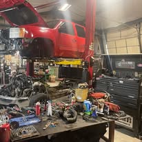 Red vehicle lifted on a hydraulic lift in a garage with tools and parts on workbenches.