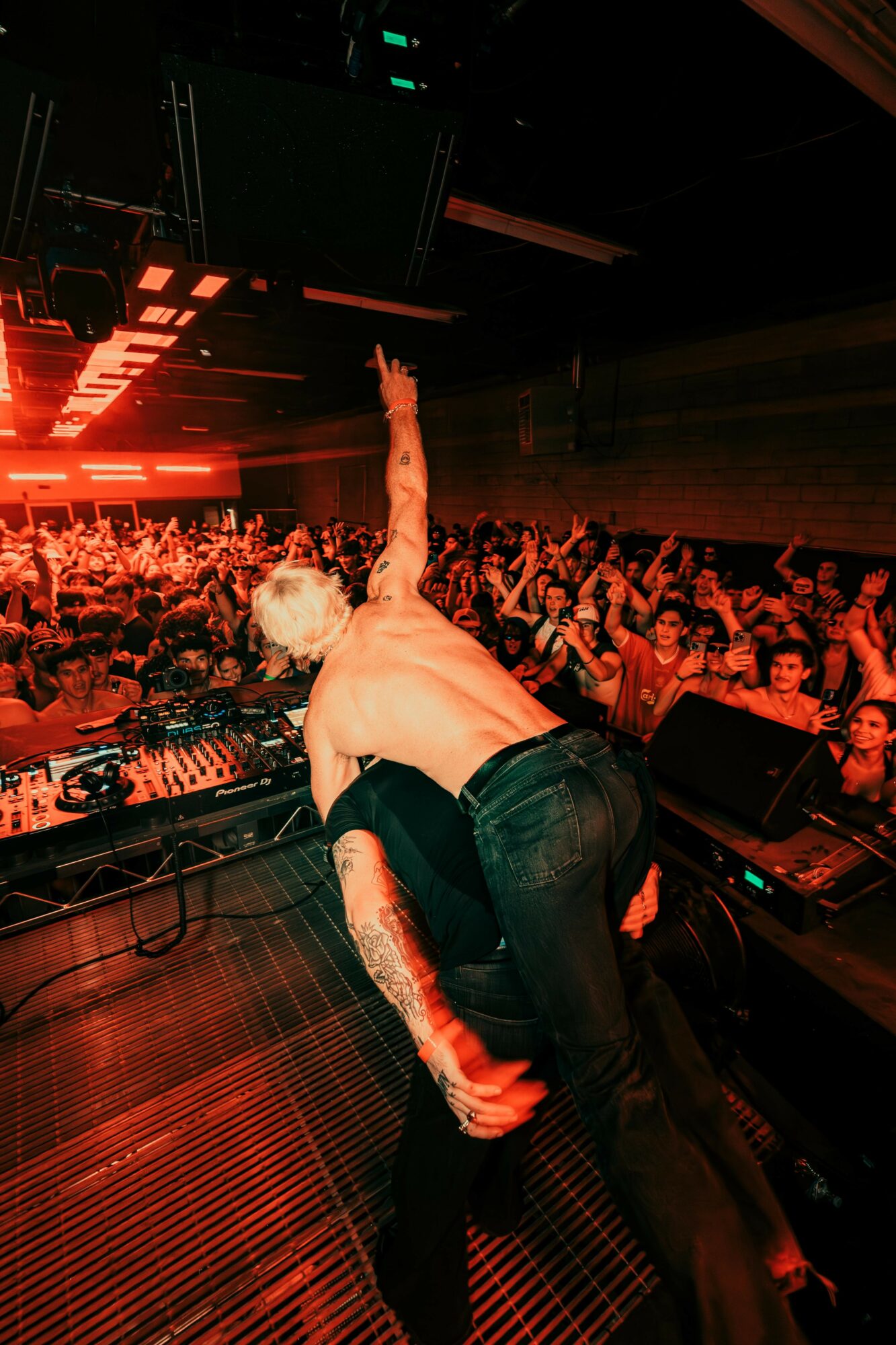 DJ performs on stage with a crowd in a dark, illuminated club setting, red lighting, and electronic equipment visible.