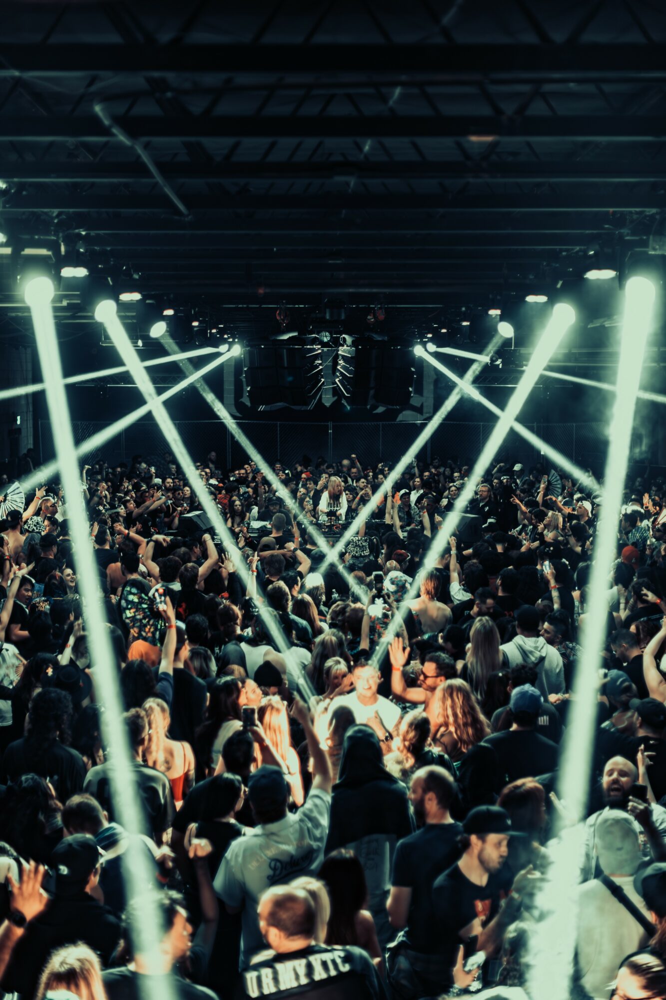Crowd at concert with bright stage lights and a dark indoor venue, people facing the stage, some with raised hands.