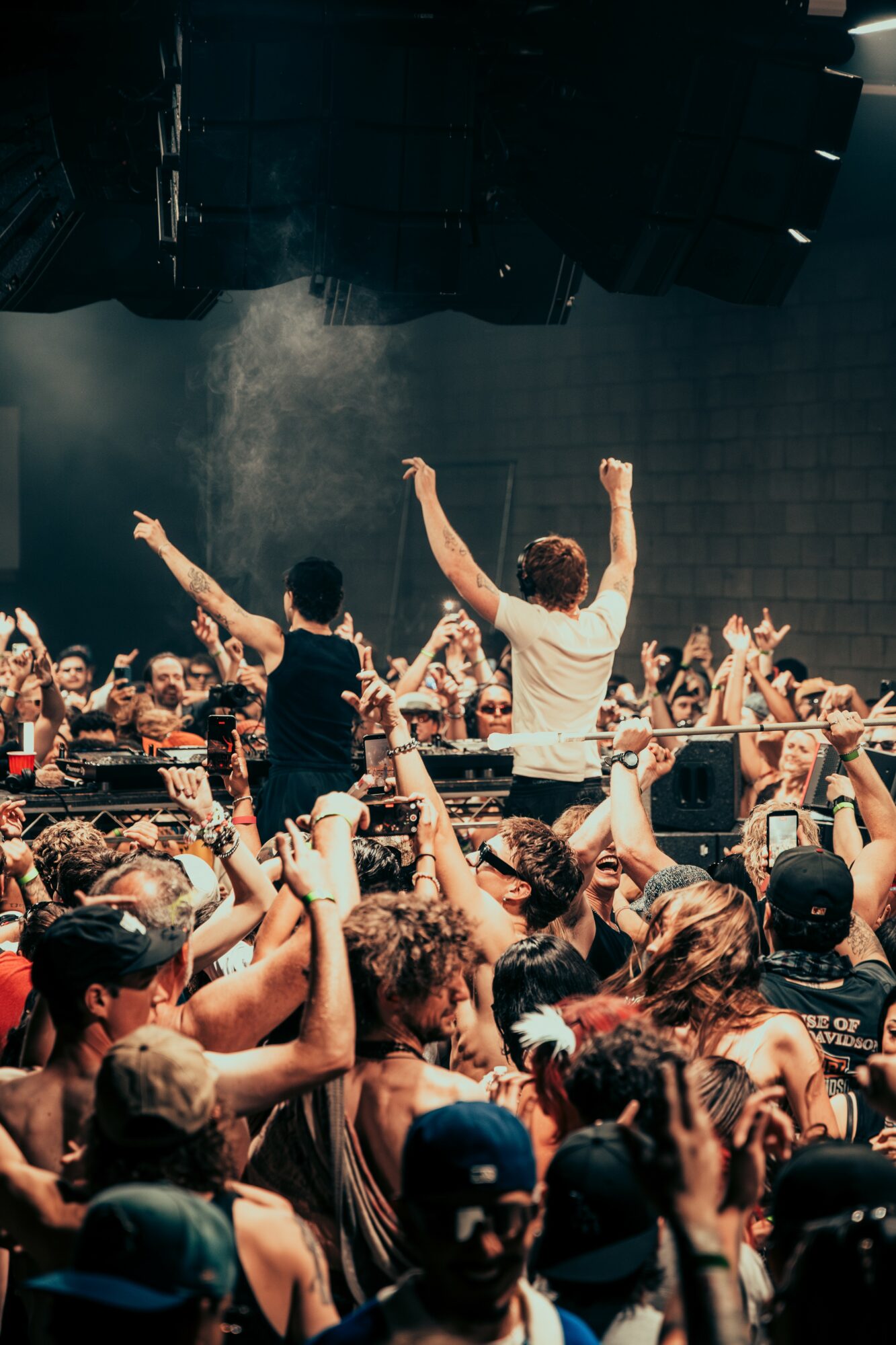 Crowd at concert with two performers on stage raising their arms, audience taking photos, dark indoor setting.