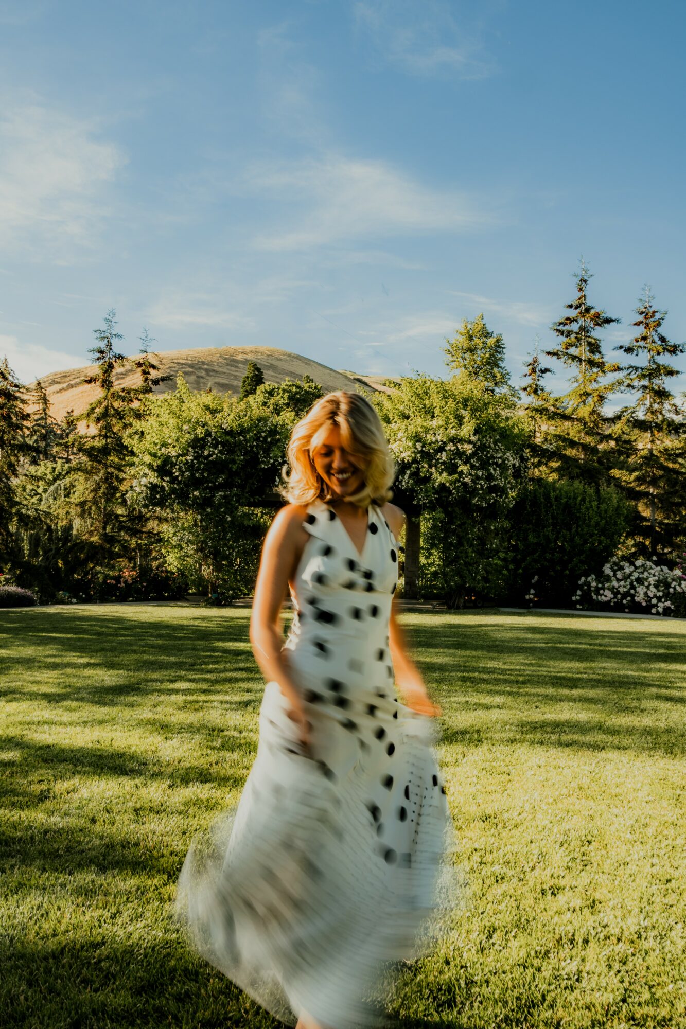 Woman in a white dress with black spots standing on grass with trees and hills in background, under blue sky.