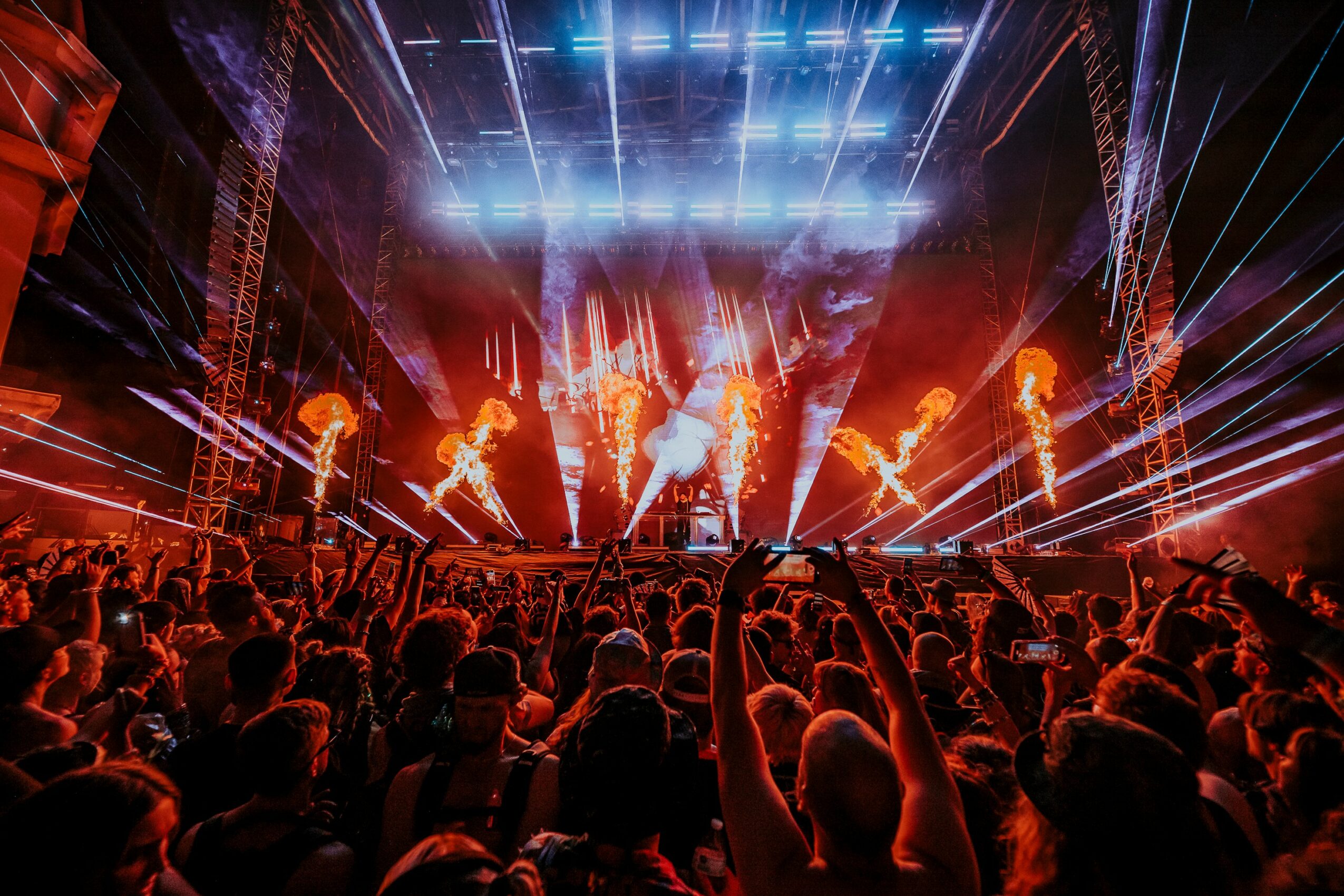 Concert scene with bright stage lights, flames, and a large crowd with raised hands.