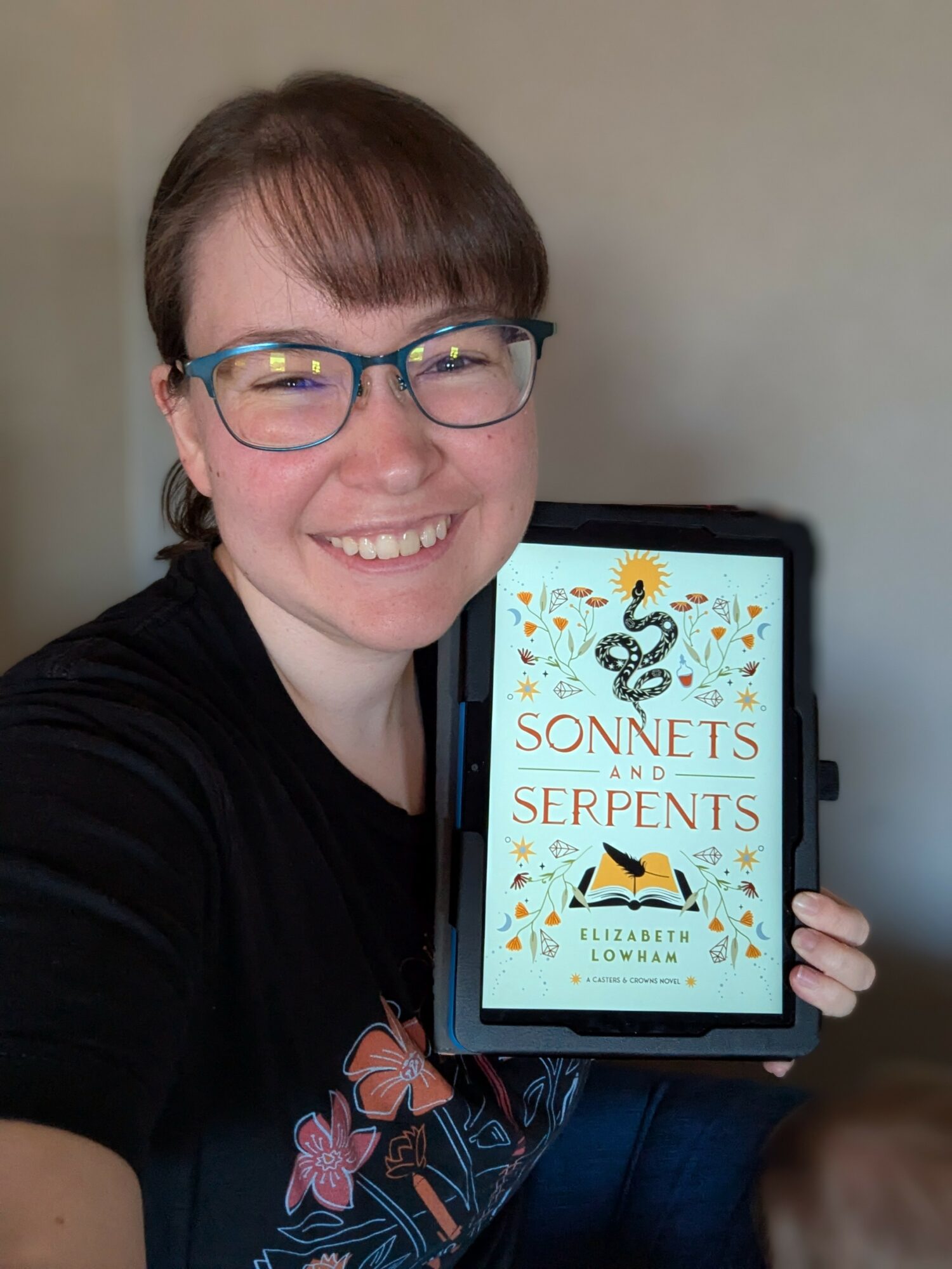 Person with glasses smiling, holding a tablet displaying a book cover titled 'Sonnets and Serpents' by Elizabeth Attaway Towarn, indoors.