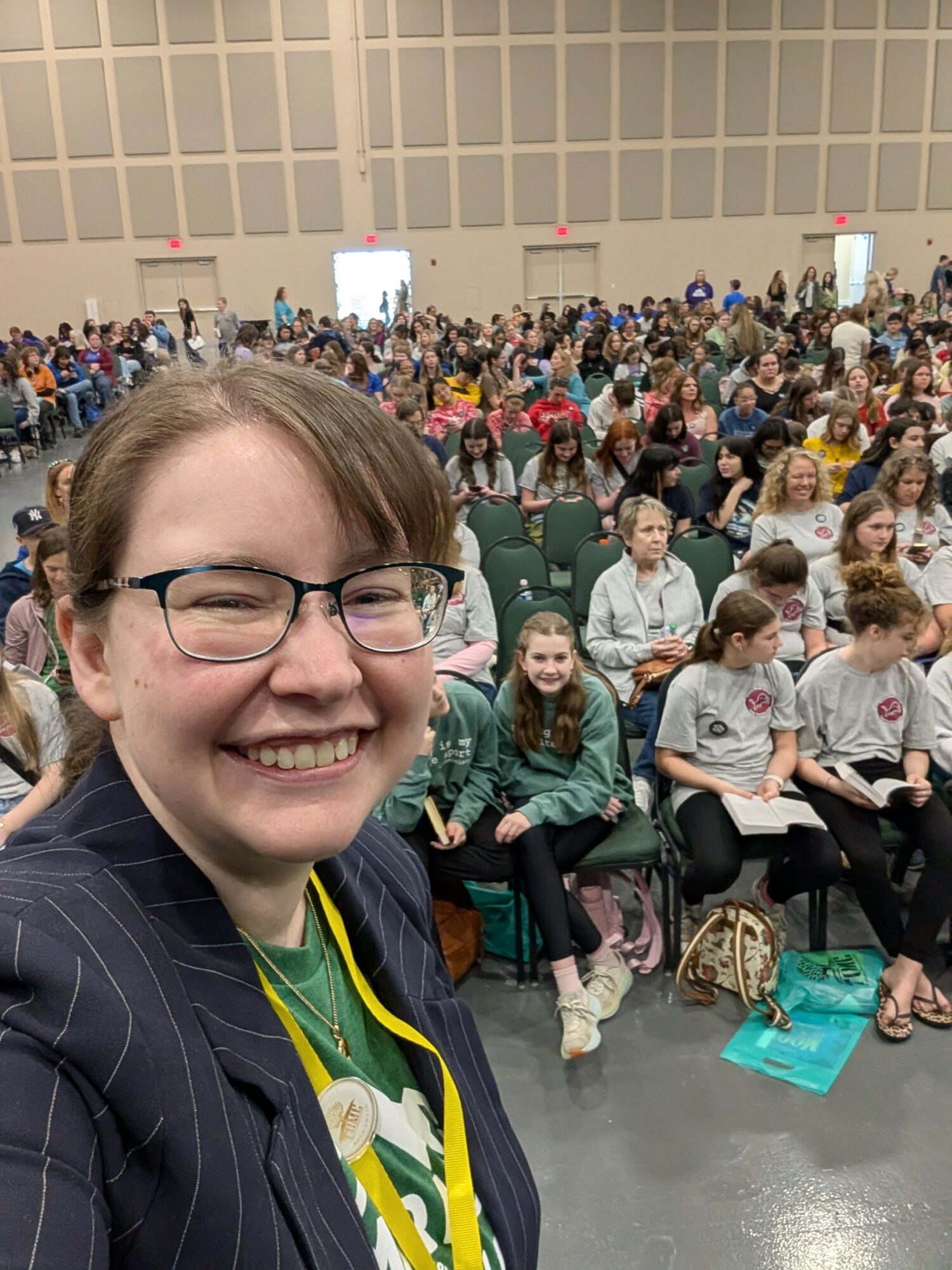 Smiling woman with glasses taking a selfie in front of a large seated audience in a spacious hall.