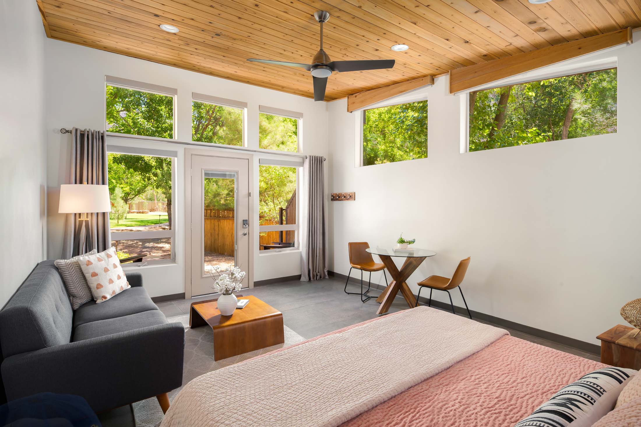 Bright living room with large windows, wooden ceiling, gray sofa, and a small dining area with two chairs.