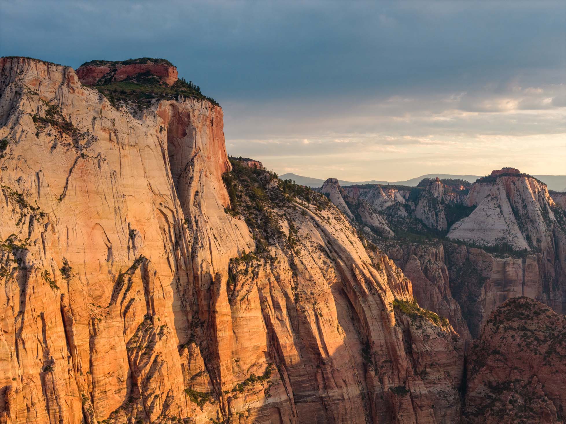 Sunlit canyon cliffs with layered rock formations and a cloudy sky in the background.