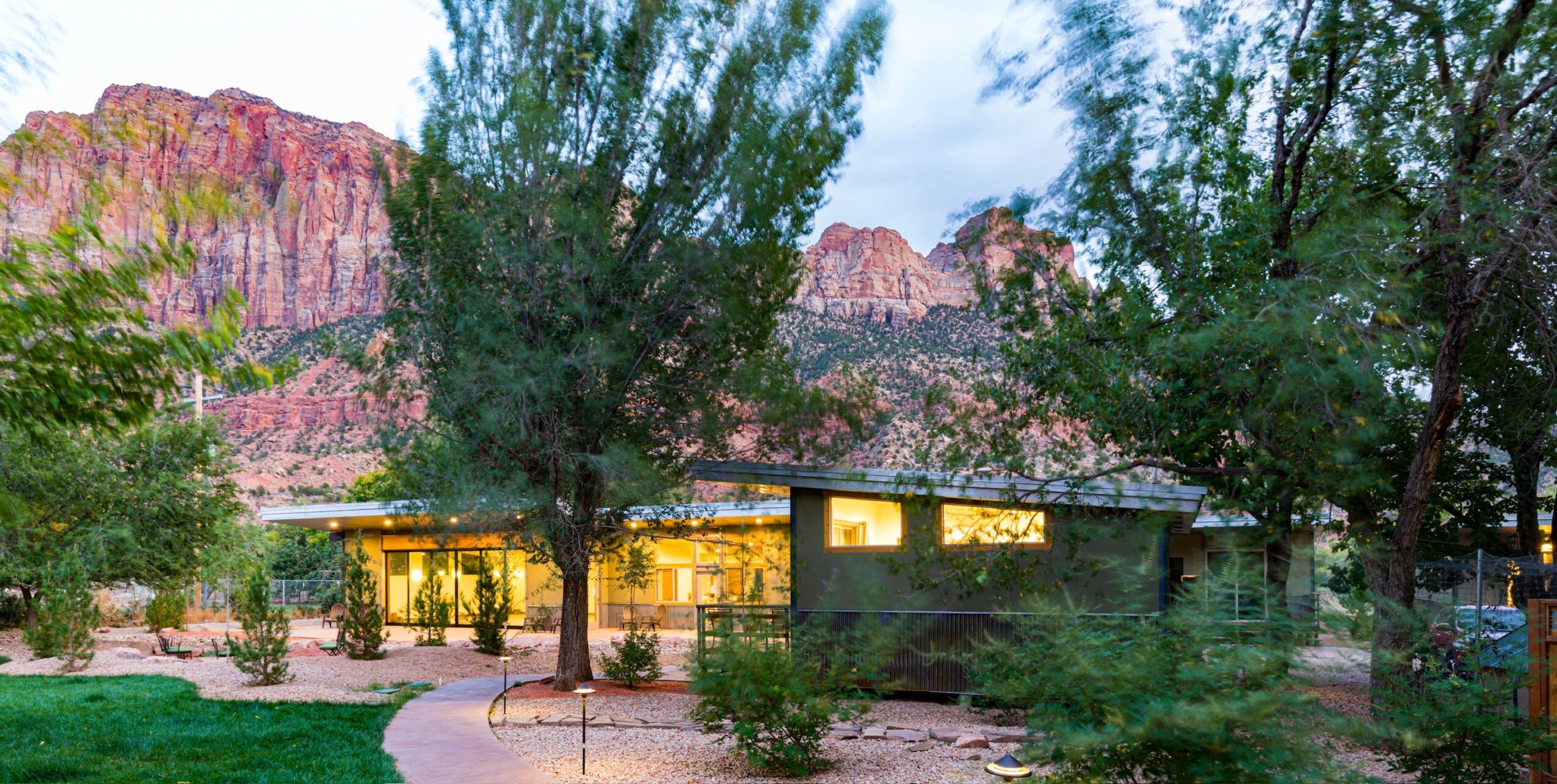 Building with large windows surrounded by trees and desert landscape with mountains in background.