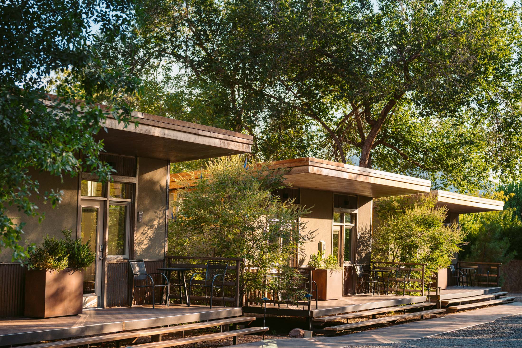 Exterior of a modern building with large windows, surrounded by trees and greenery, with a small outdoor seating area.