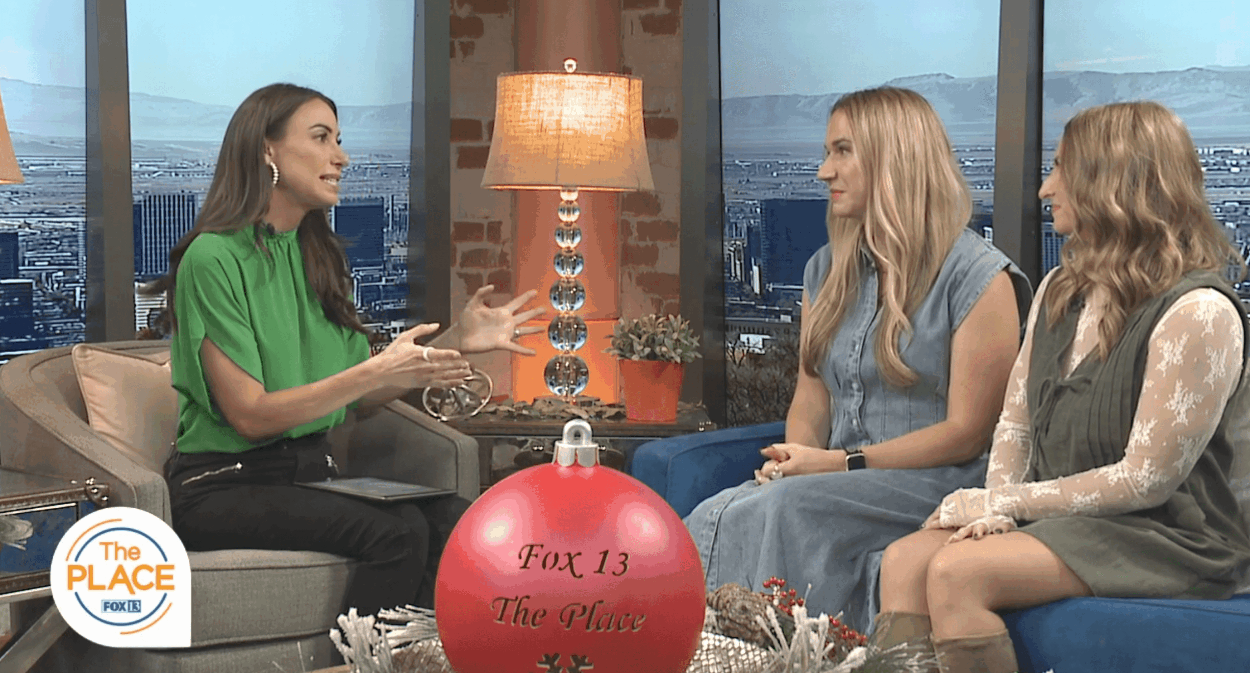 Three women sit on a TV set with a city view background, engaged in conversation, with a large red ornament in front.