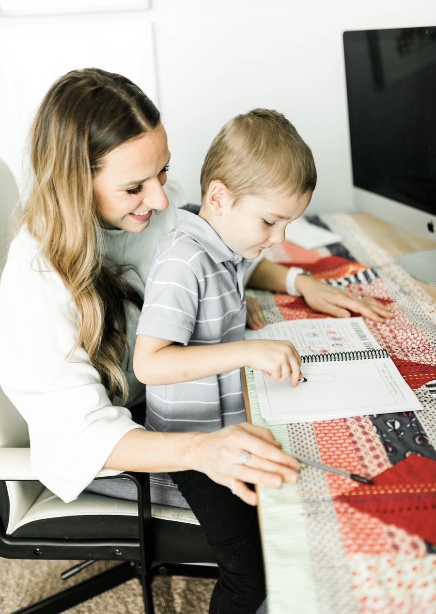 Woman and young boy sitting at a table, looking at a notebook, with a computer monitor in the background.