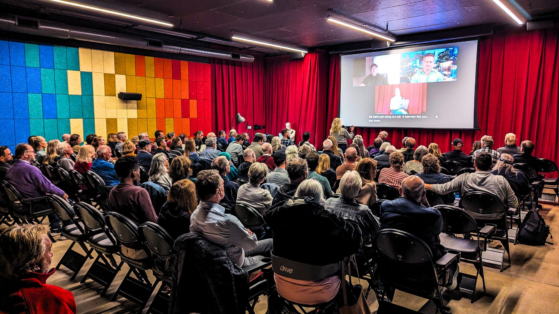 Audience seated in a conference room watching a presentation on a large screen, with colorful wall panels and red curtains.