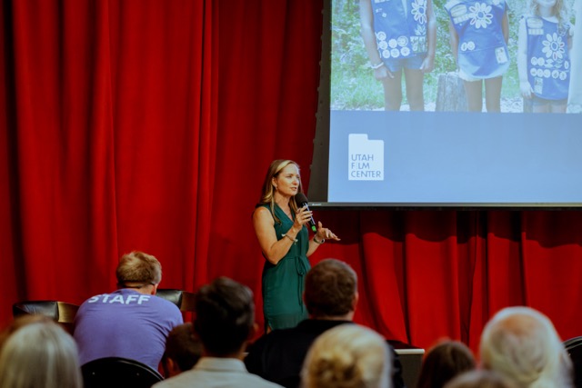 Woman in green dress speaking on stage with red curtain background, audience seated, large screen displaying a photo, and a logo.
