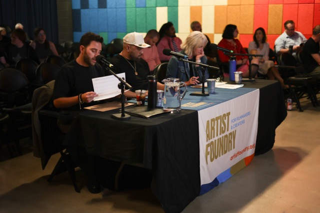 Panel of four people seated at a table with microphones, papers, and water bottles, in front of an audience, at an event called Artist Fluency.