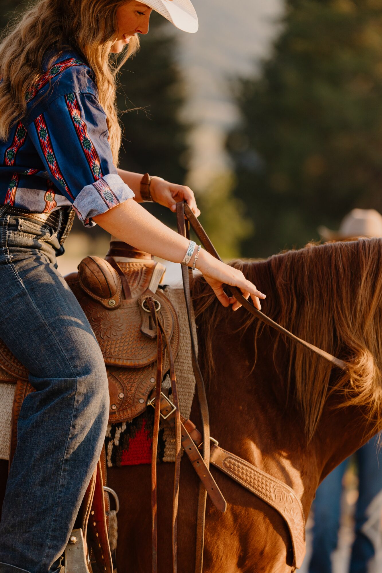 Young woman in a cowboy hat and plaid shirt riding a horse outdoors, pointing forward, with trees in the background.