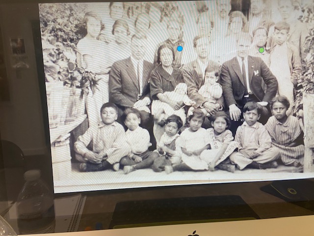 Group of children and adults sitting and standing outdoors, some children sitting on the ground, in black and white.