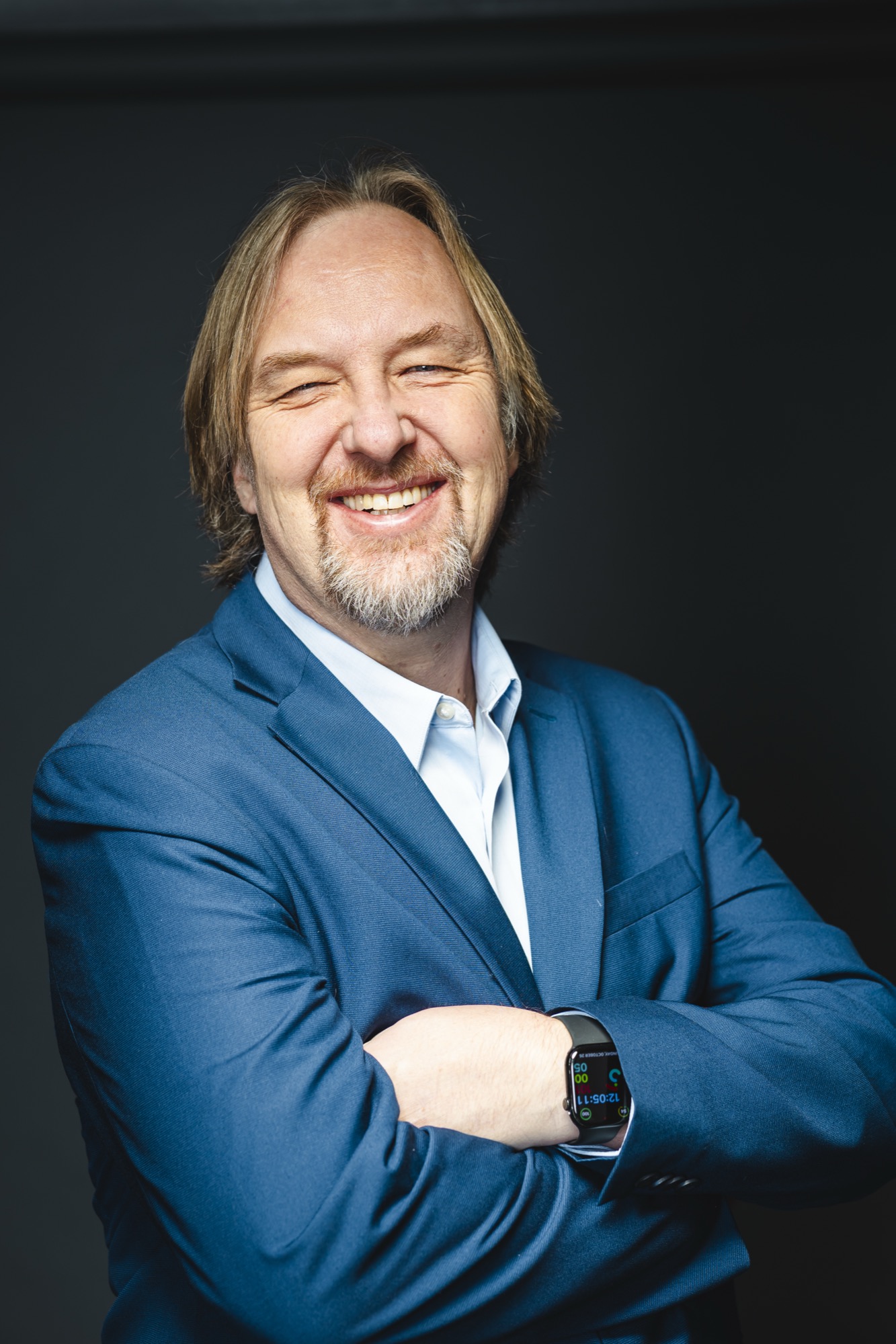 Smiling man with shoulder-length hair and beard, wearing a blue suit and white shirt, standing with arms crossed against a dark background.