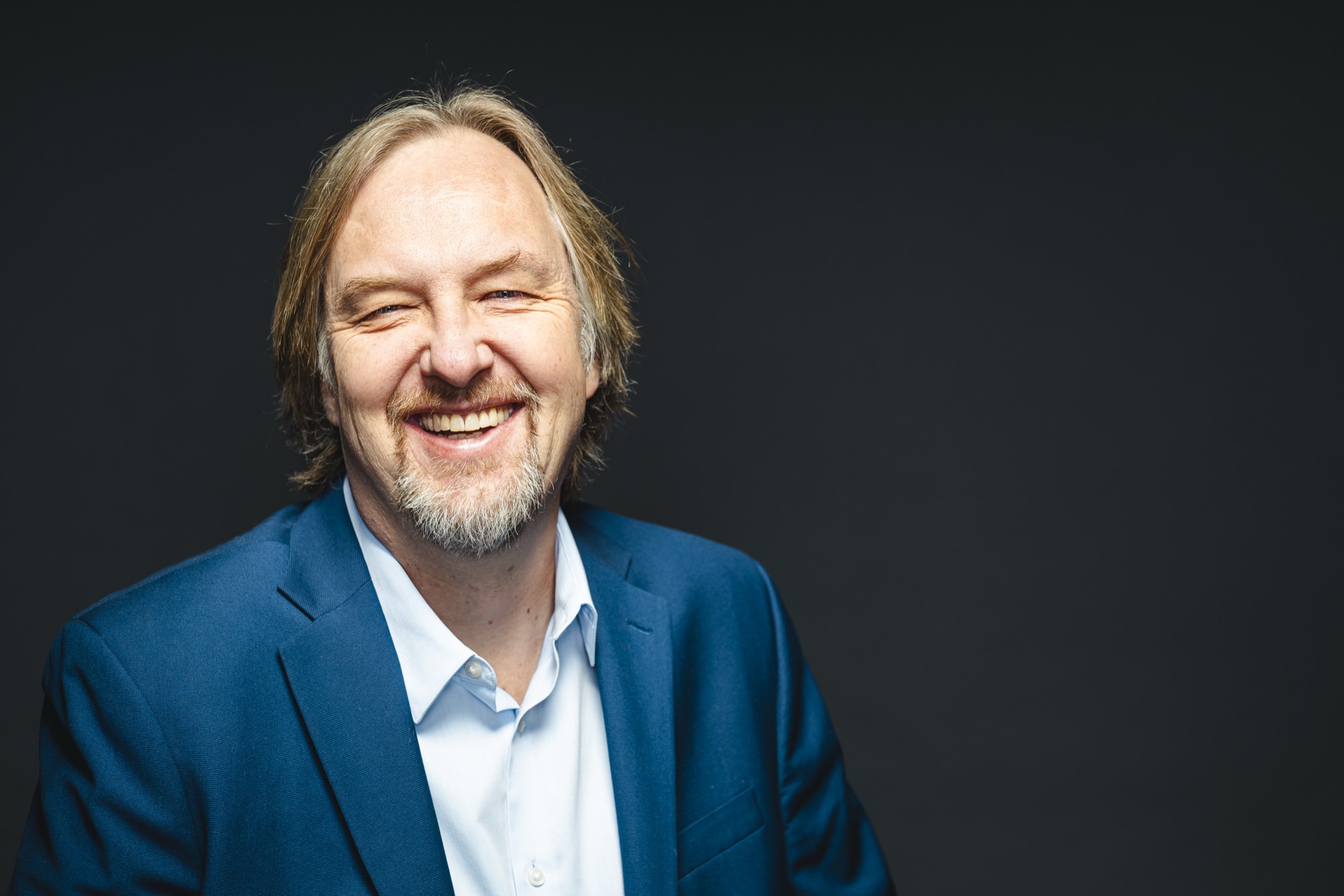 Smiling man with shoulder-length hair and beard, wearing a blue suit and white shirt, against dark background.