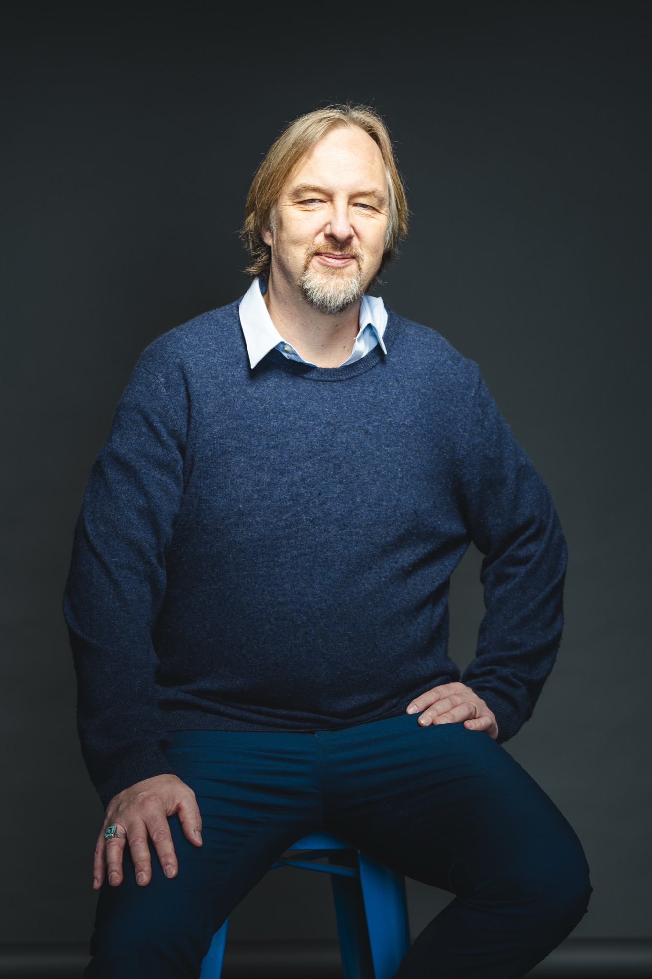 A middle-aged man with shoulder-length hair and a beard, wearing a blue sweater and white shirt, sitting on a stool against a dark background.