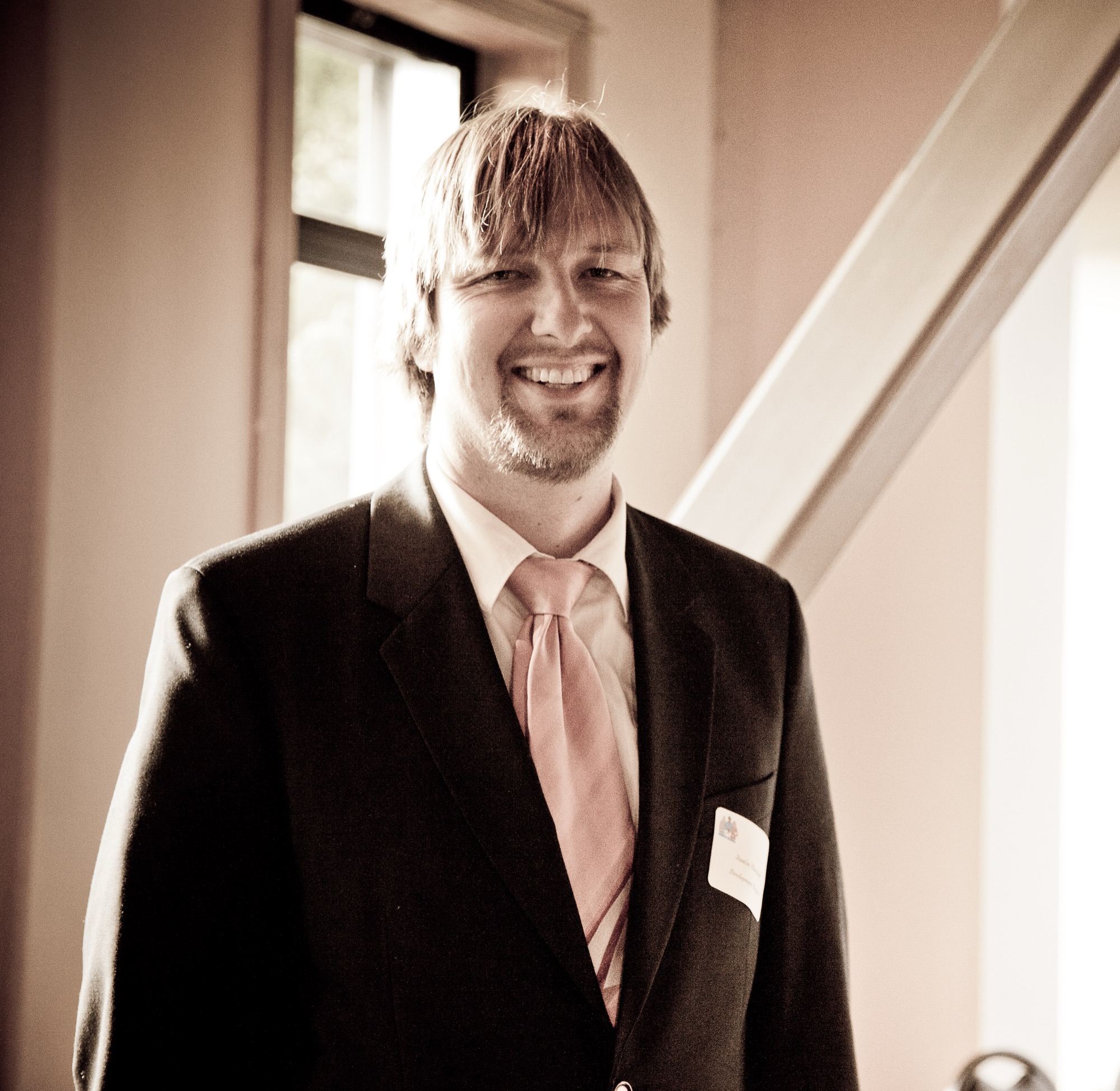 Smiling man in a suit and tie standing indoors near a window with natural light.