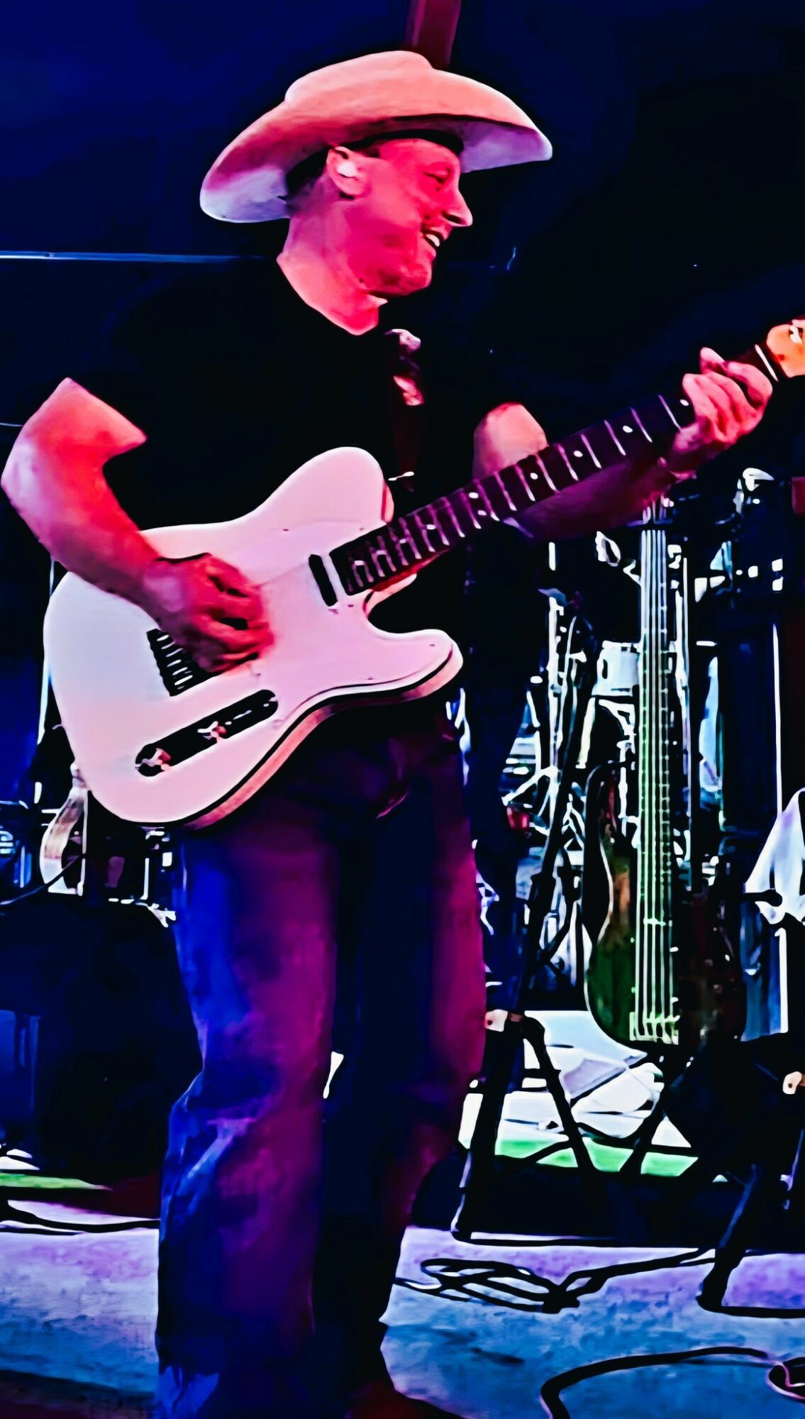 Person wearing a cowboy hat playing an electric guitar on stage, with musical equipment in the background.