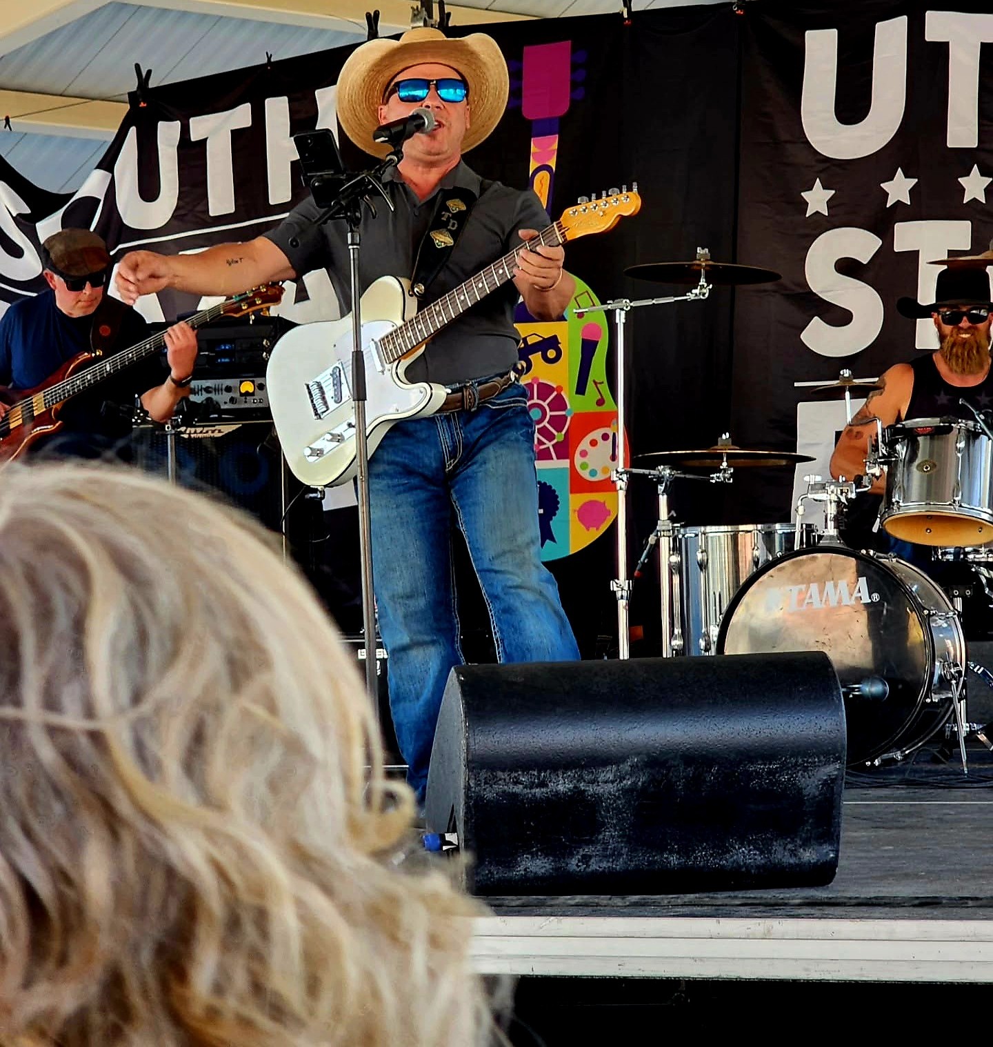 Musicians perform on stage with a banner behind them, one wearing a cowboy hat and sunglasses, playing guitar and drums.