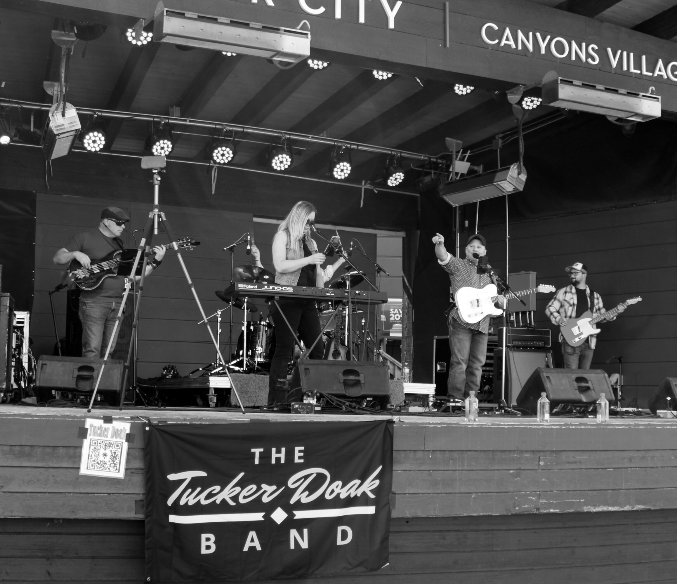 Band performs on stage with five musicians, instruments, and a banner reading 'The Tucker Doak Band'.