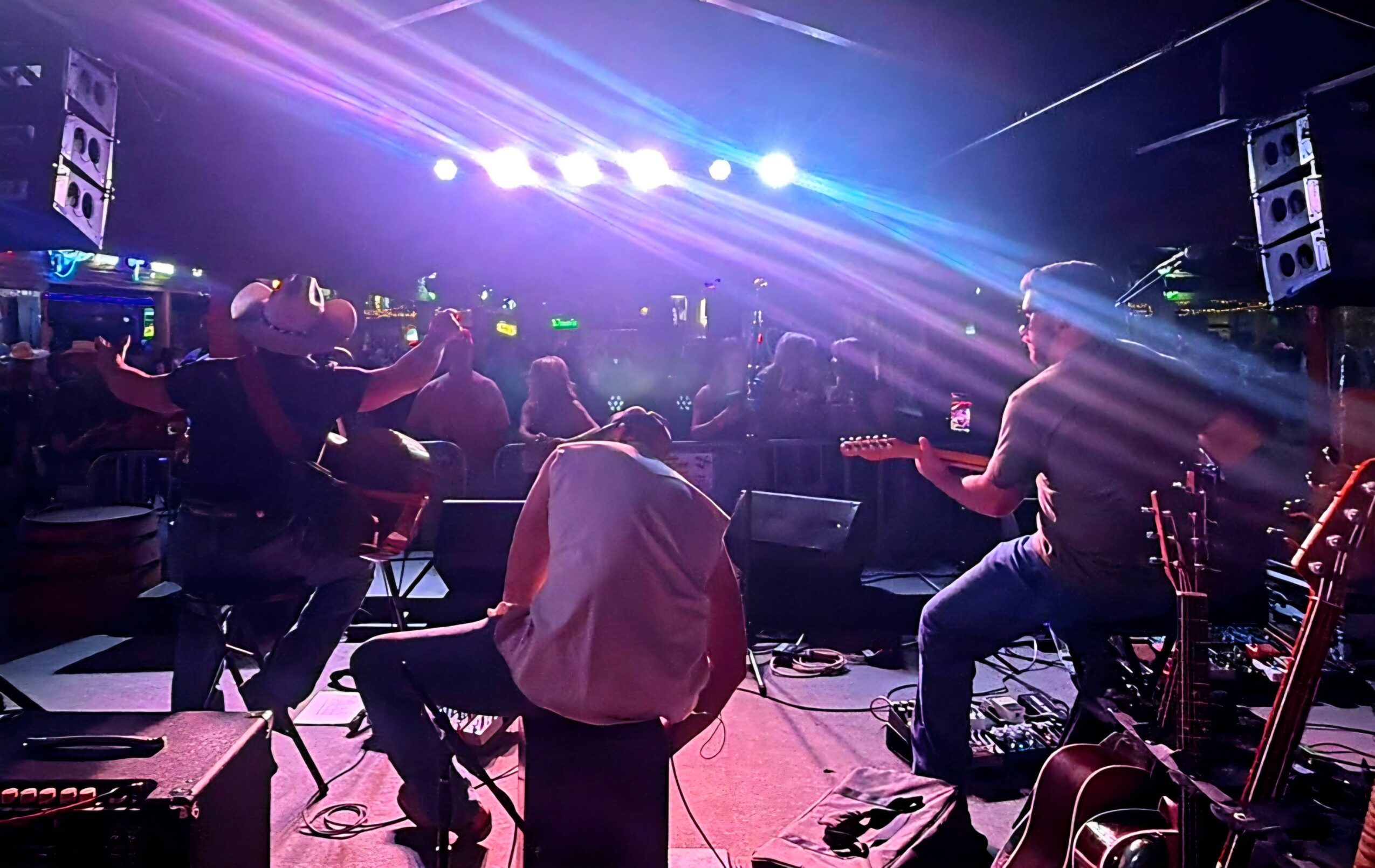 Musicians perform on stage with colorful lights, audience watching in a dark venue, guitars and equipment visible.