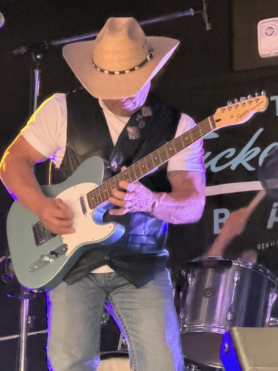 Person wearing a cowboy hat playing an electric guitar on stage, with drums in the background.