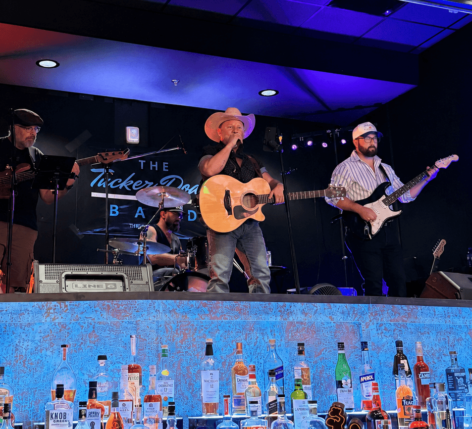 Four musicians perform on stage with instruments, a drum set, and microphones, under blue and purple stage lighting.