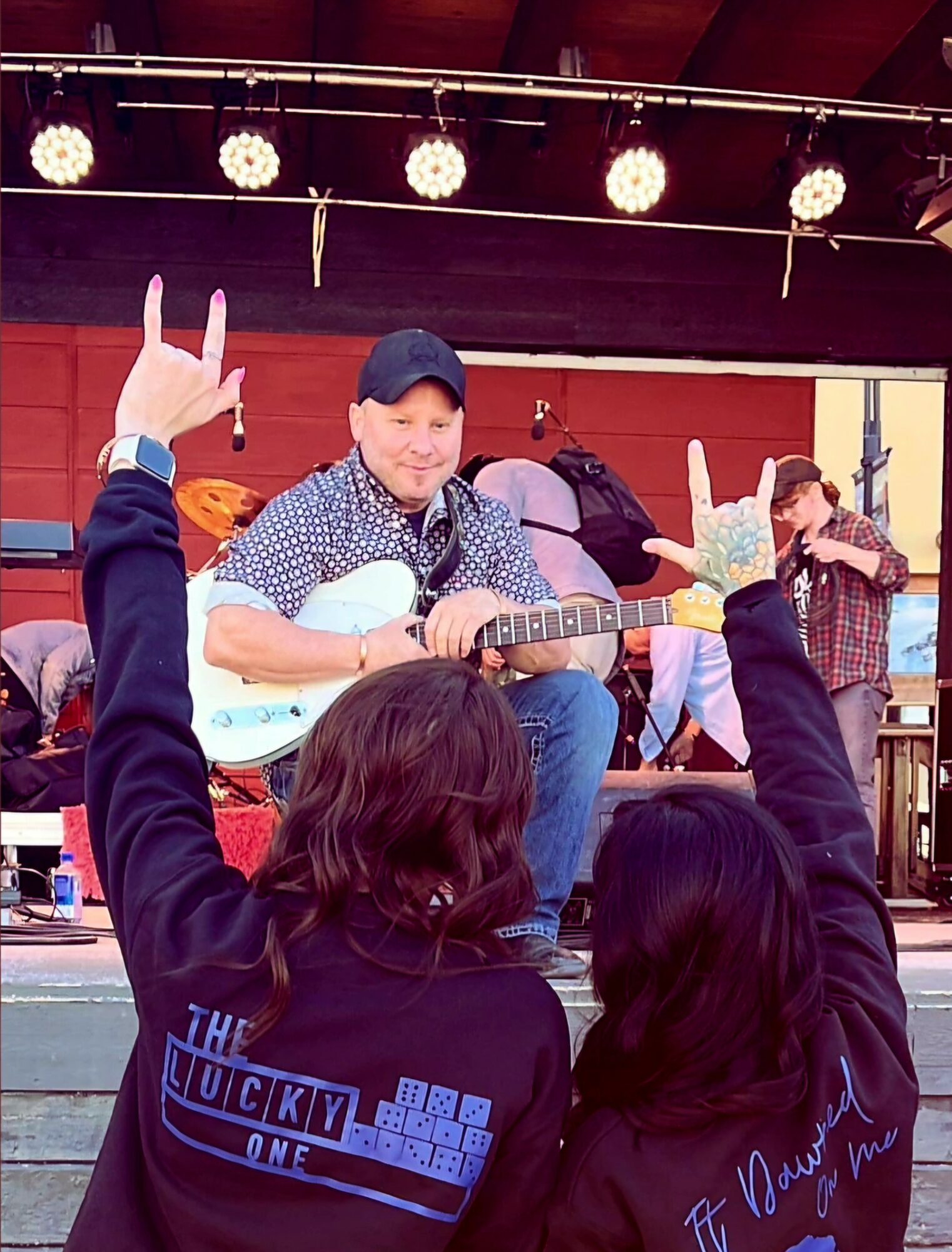 Person with long hair and a black hoodie with blue text and graphics, raising hands with peace signs, watching a young man playing guitar on stage.