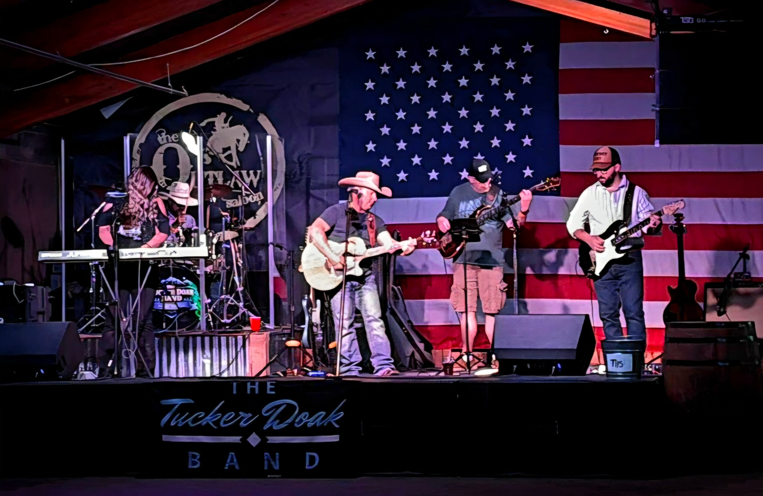Five musicians perform on stage with American flag backdrop, playing guitars and singing, with band name displayed below.