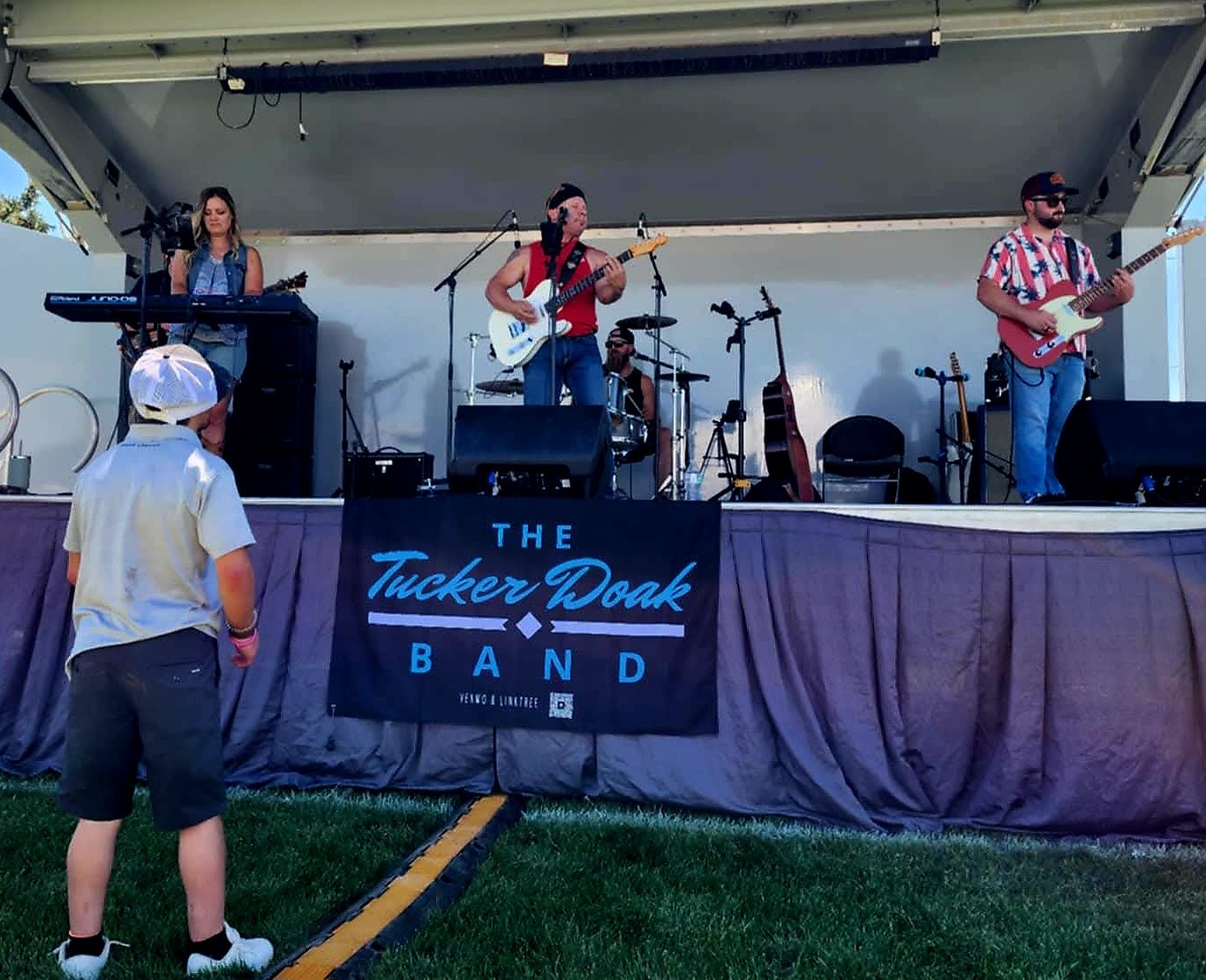 Band performing on stage with a keyboard, guitar, and drums, and a sign reading 'The Tucker Deak Band.' Two children watch.
