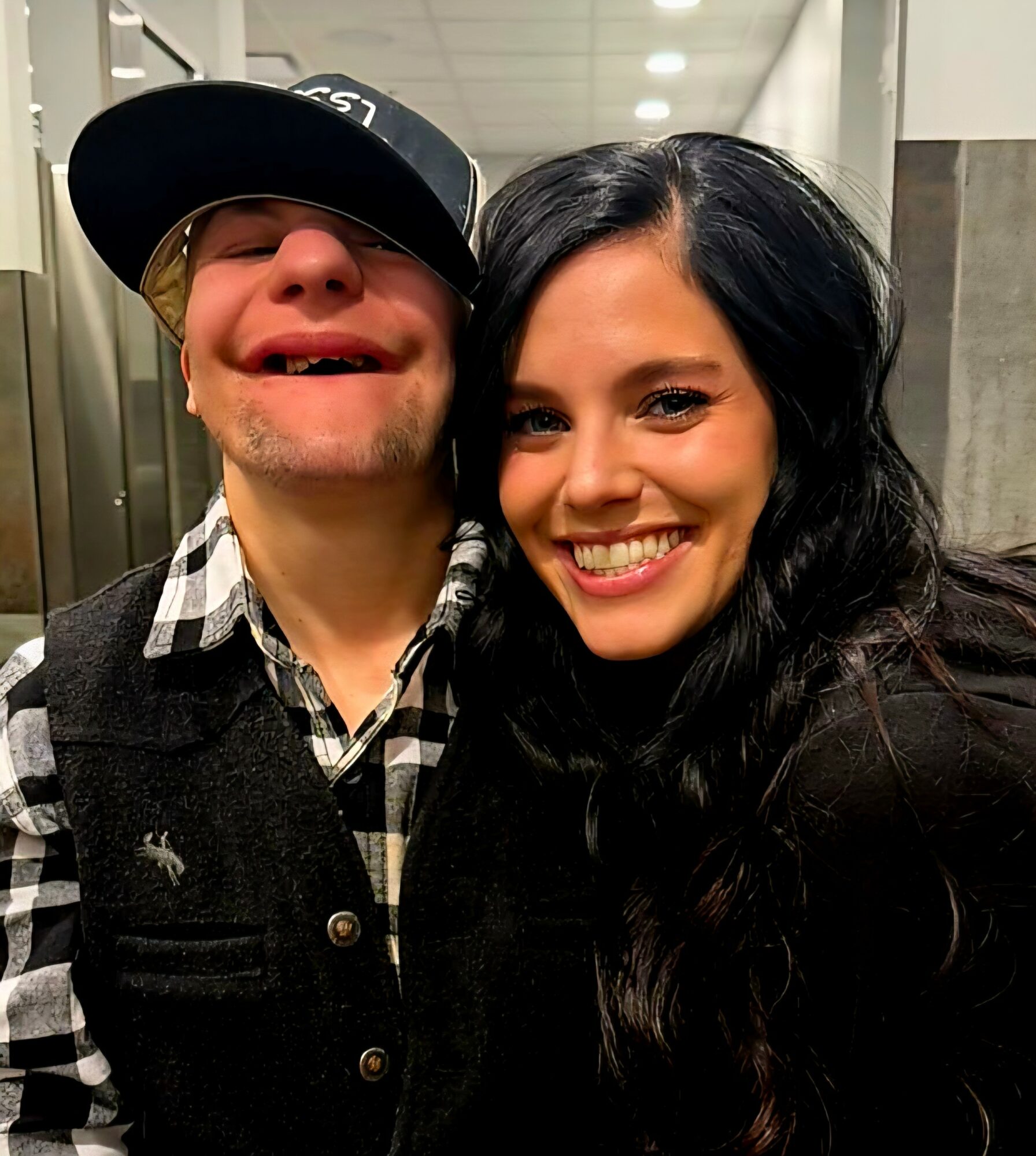 Two people smiling, one wearing a hat and glasses, the other with long dark hair, in an indoor setting.