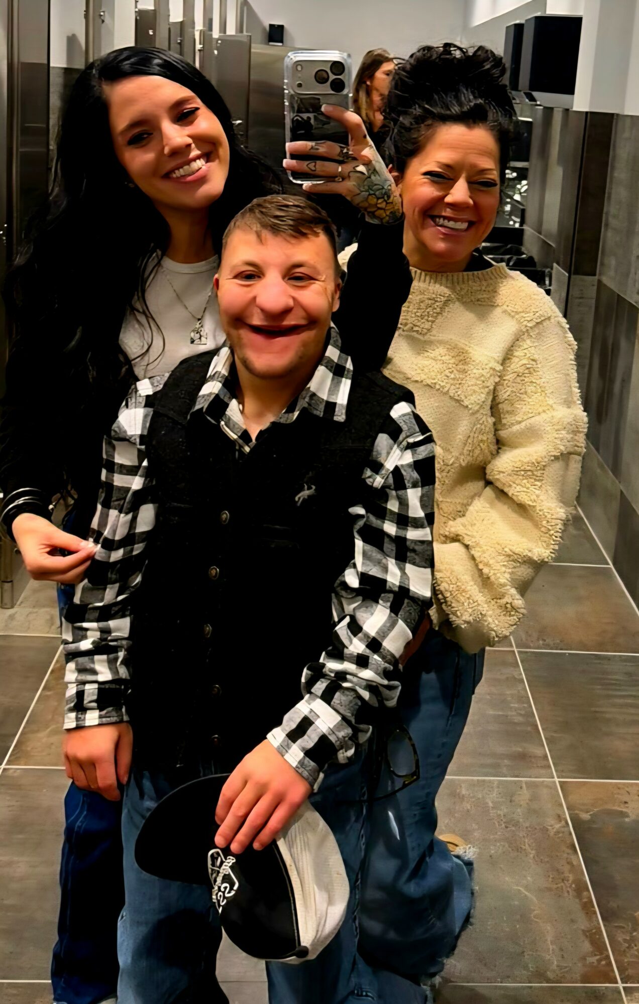 Three people smiling in a mirror selfie, two women and a young boy, in a tiled room.